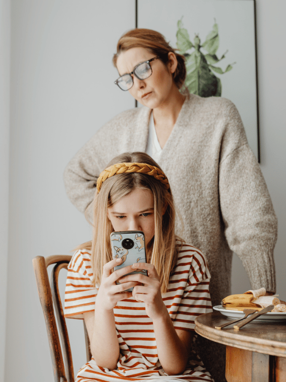 Parent standing over a young girl on her smartphone, questioning age of smartphone use.
