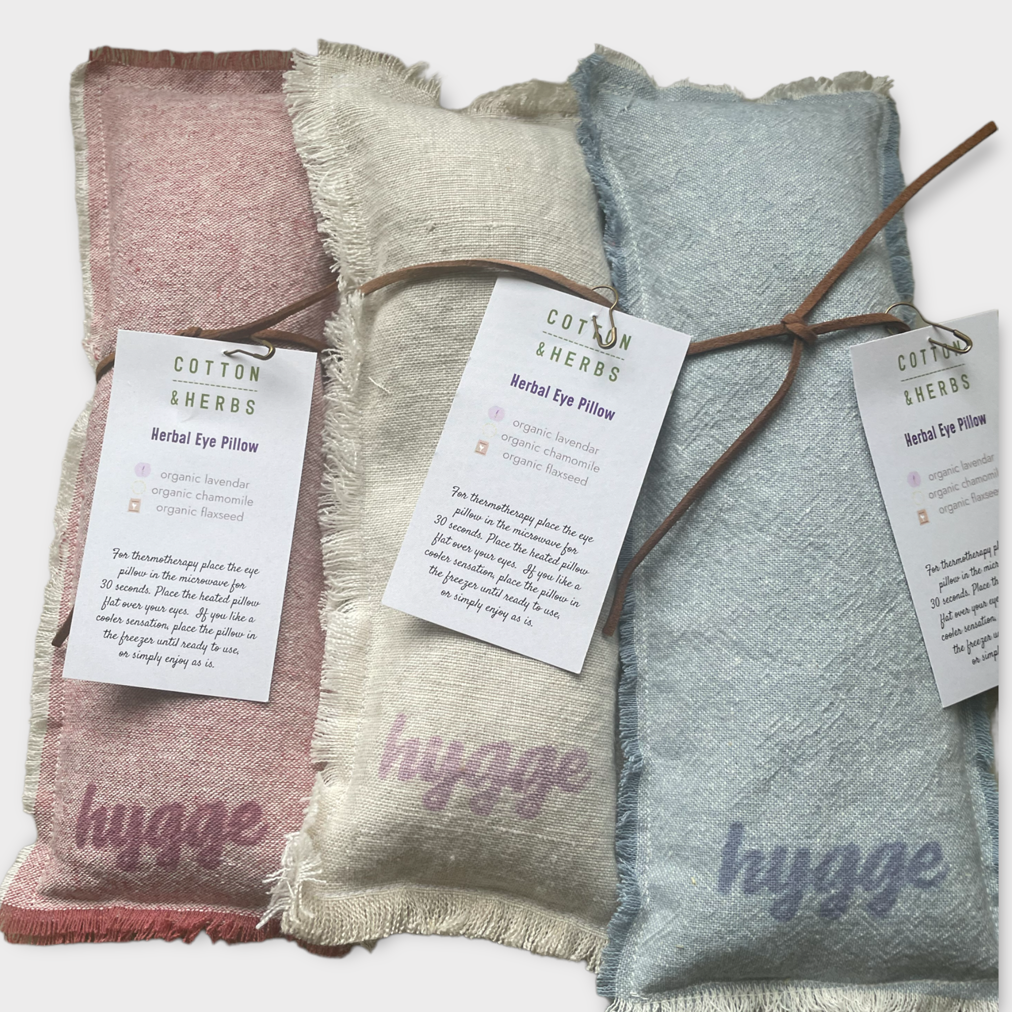 Three cotton and herbs herbal eye pillows in pink, beige, and blue, each with a tag providing ingredients and usage instructions.