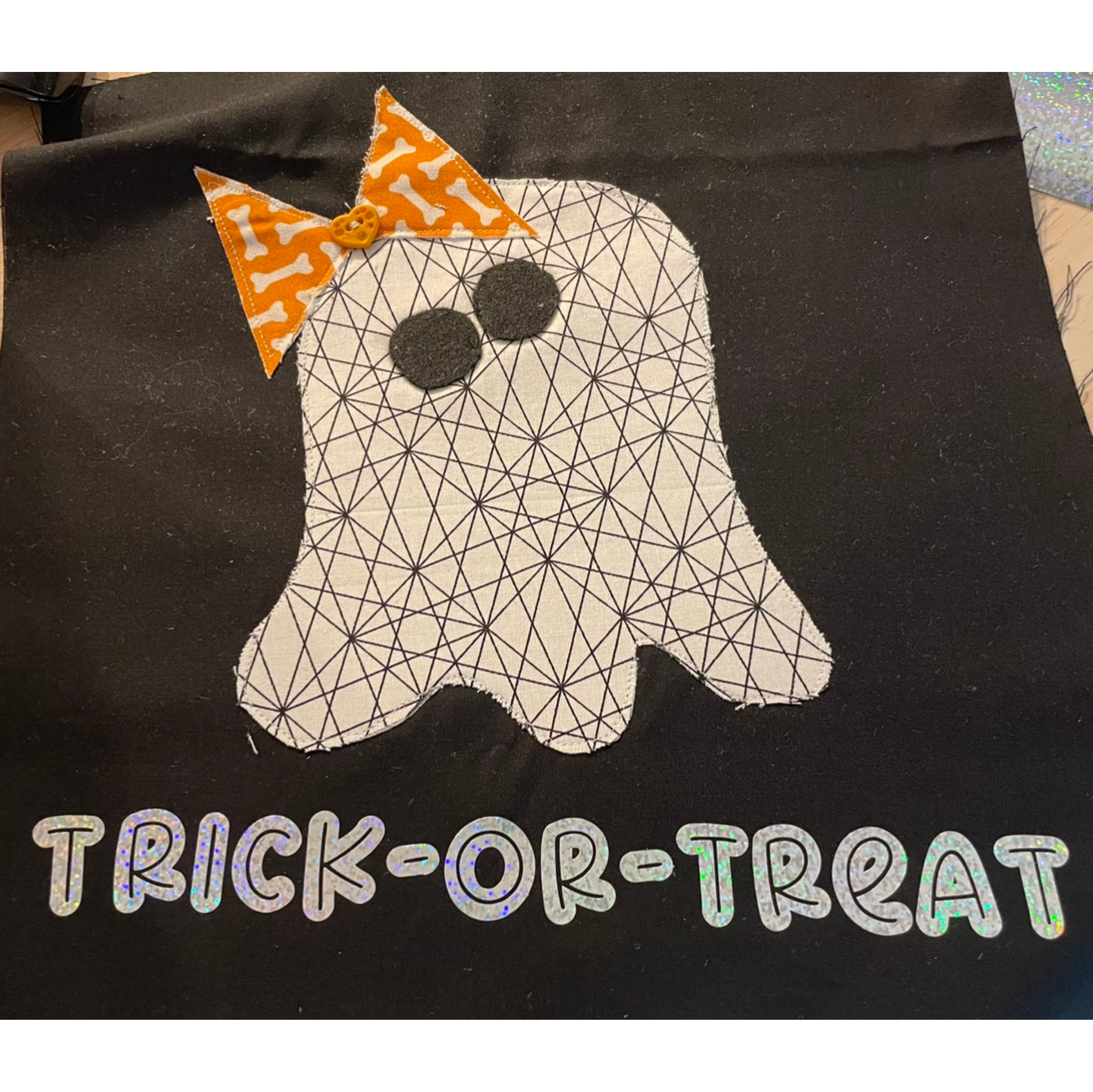 A Halloween-themed fabric decoration featuring a ghost shape with a geometric pattern, garnished with an orange bow with white bones and a small orange button. The background is black, with holographic text beneath that reads 'TRICK-OR-TREAT'.
