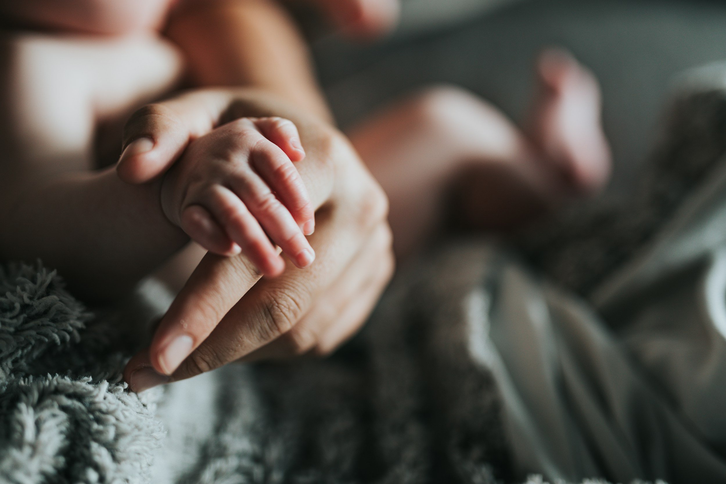 A close-up of a small baby's hand holding an adult's finger, with the baby's body slightly out of focus in the background.