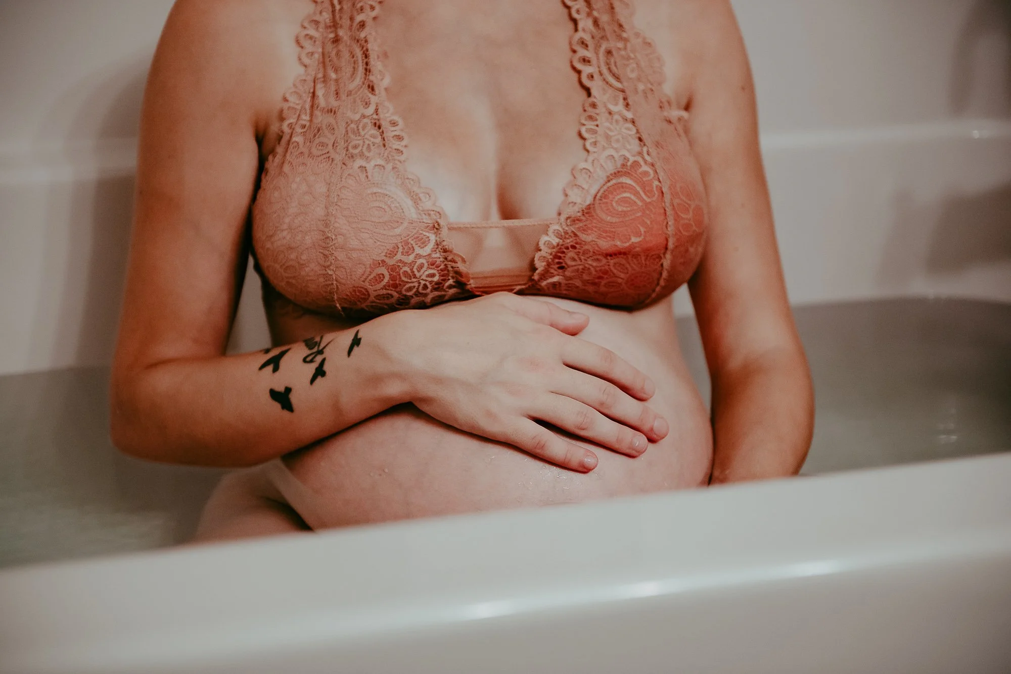 A woman wearing a lace bra is sitting in a bathtub, holding her pregnant belly with one hand, with a tattoo of birds on her arm.