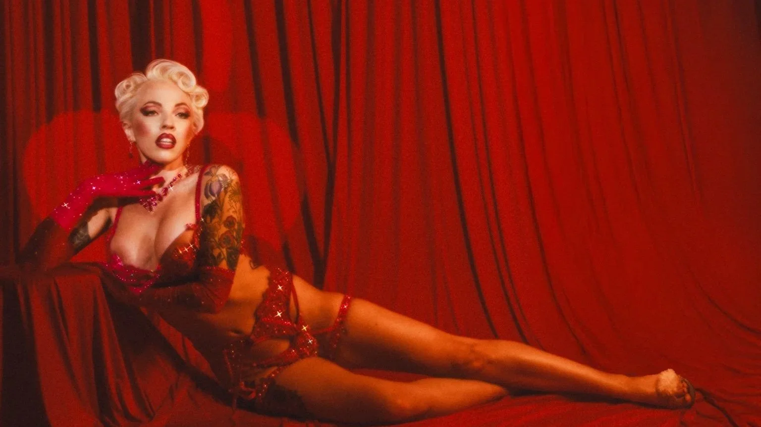 A blonde woman with vintage hairstyle lying on red velvet curtains, wearing red lingerie with glitter and long red gloves, posing seductively.