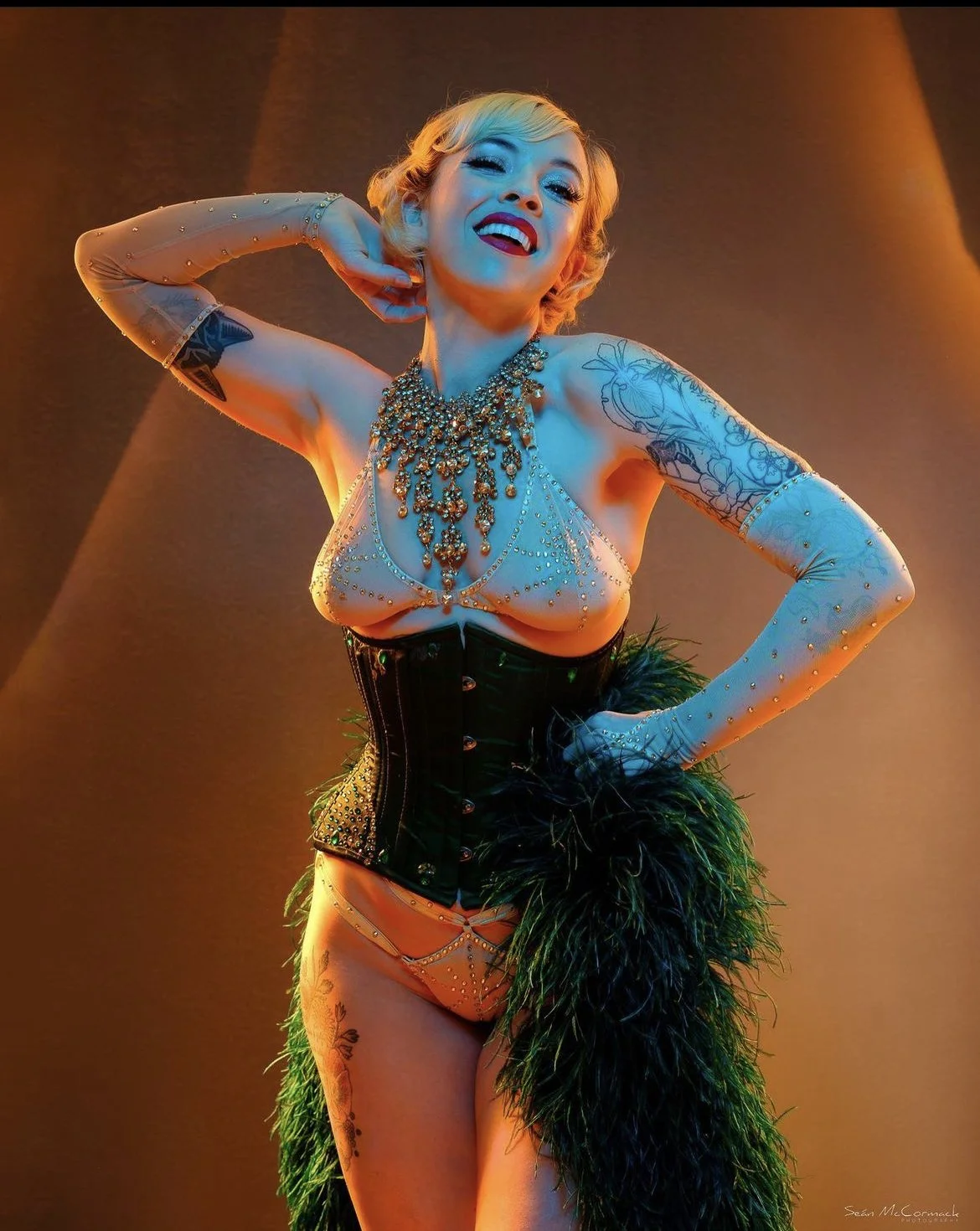 A woman with blonde hair posing confidently, wearing a jeweled top, corset, gloves, and a feathered skirt, with a warm brown background.
