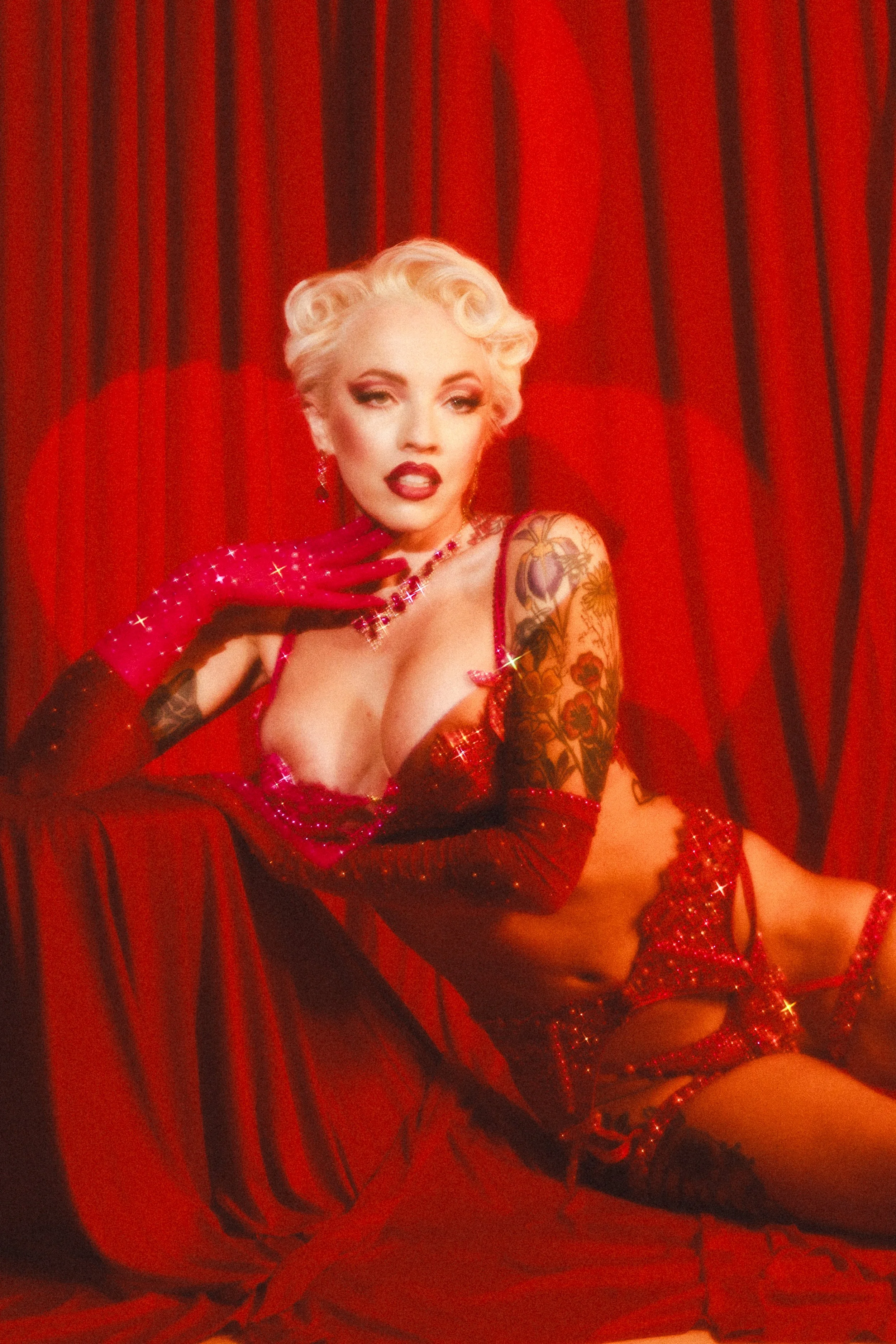 A woman with platinum blonde hair styled in vintage curls, wearing red sequined gloves, red lingerie, and thigh-high stockings with lace tops, posing in front of red curtains.