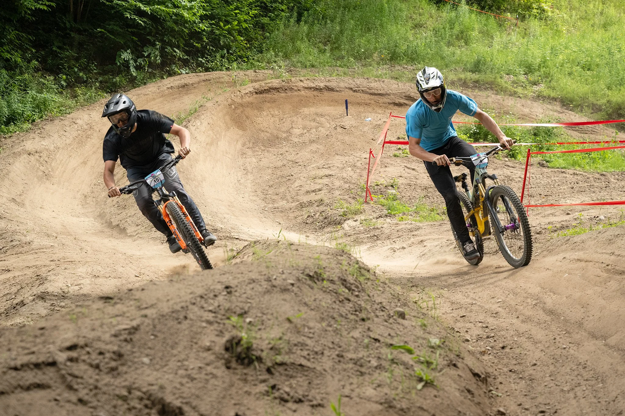 Two mountain bikers racing on a dirt track with curves, surrounded by greenery.