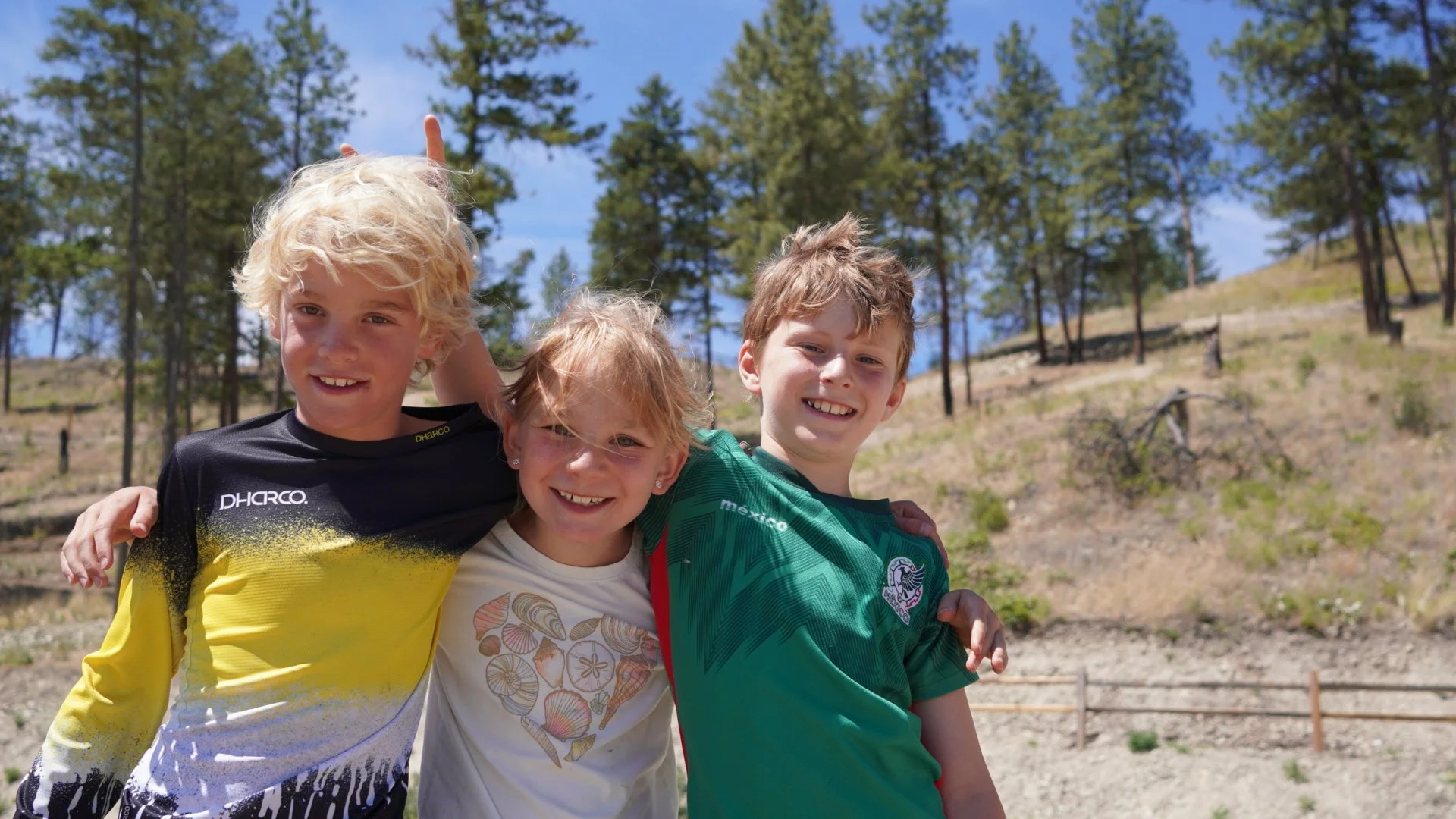 Four children smiling and hugging outdoors in a wooded area with trees and a fenced path in the background.