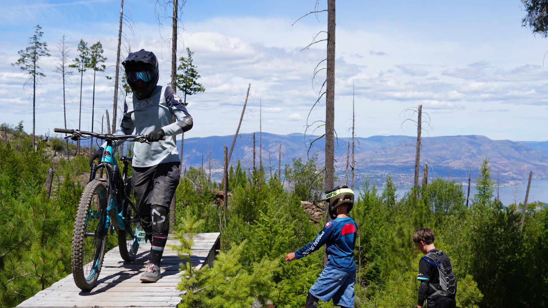 Children and an adult mountain biking on a trail through a forest with distant mountains and lake amenities.