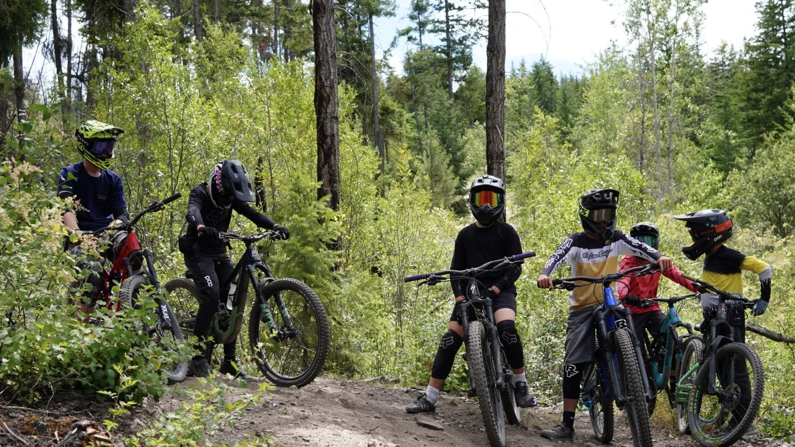 Group of children in mountain biking gear with helmets and goggles, standing with bikes on a trail in a forested area.