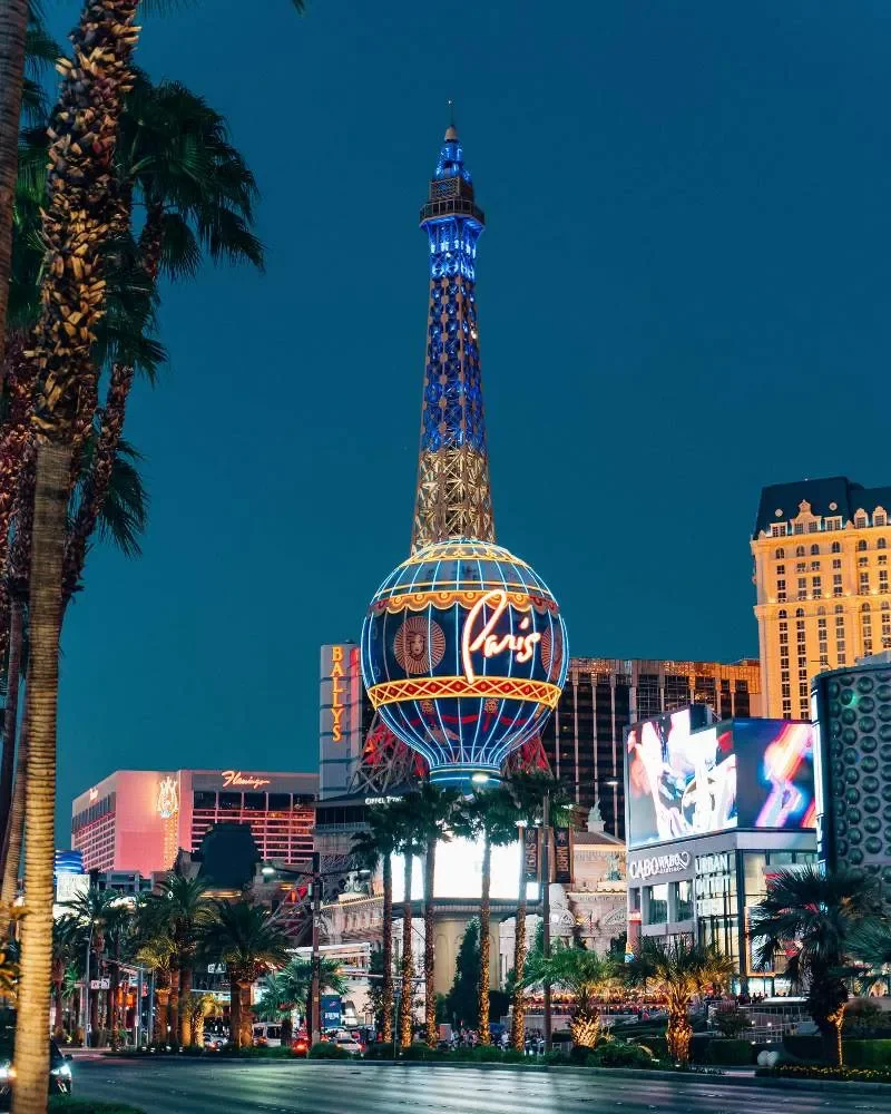 Night view of the Las Vegas Strip featuring the replica of the Eiffel Tower with neon lights, palm trees, billboards, and casino signs.