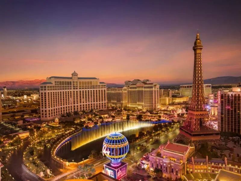 Night view of Las Vegas Strip with hotels, the Eiffel Tower replica at Paris Las Vegas, and the Bellagio fountain with colorful lights.
