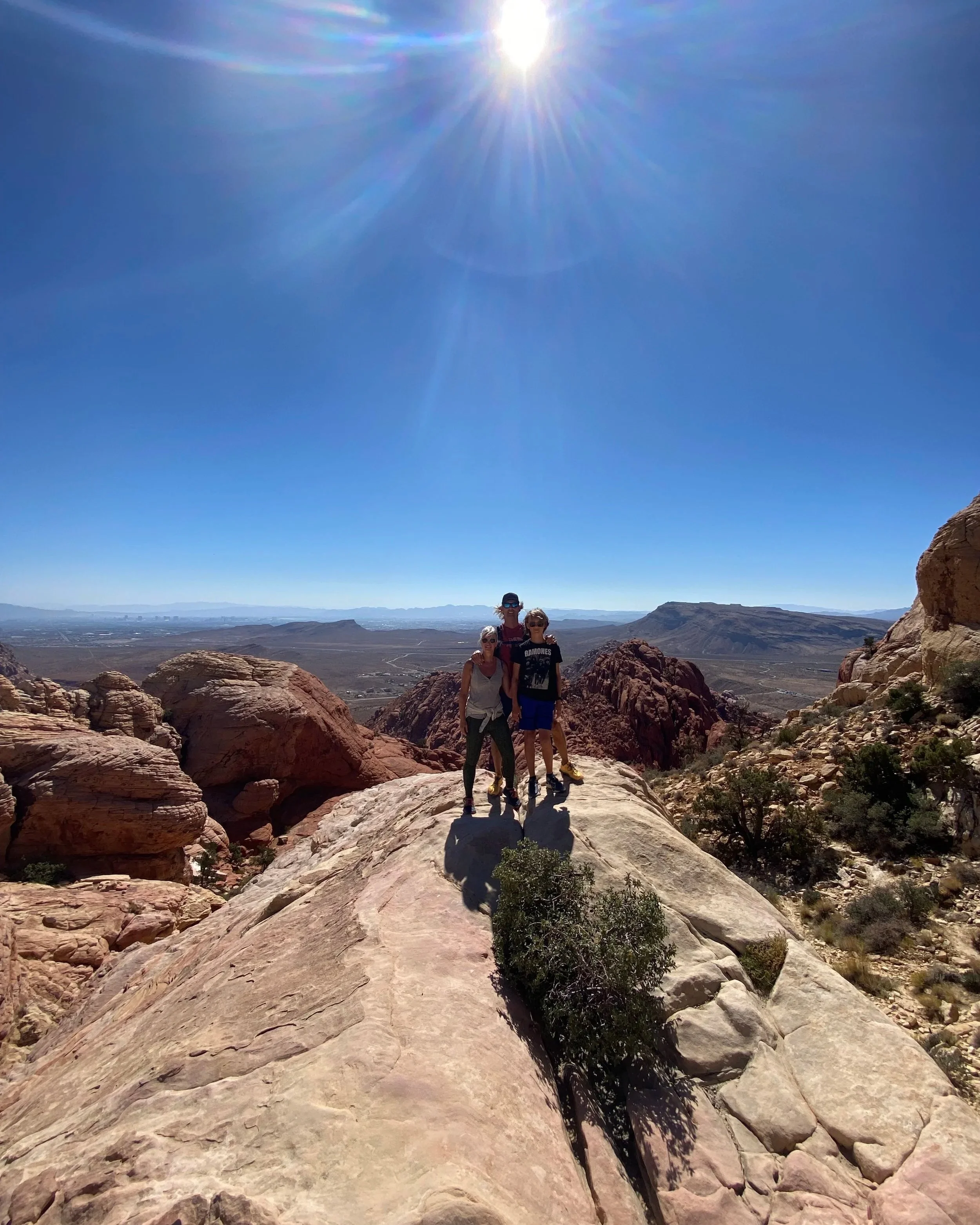 Three people standing on a rocky outcrop in a desert landscape under a bright sun and blue sky.