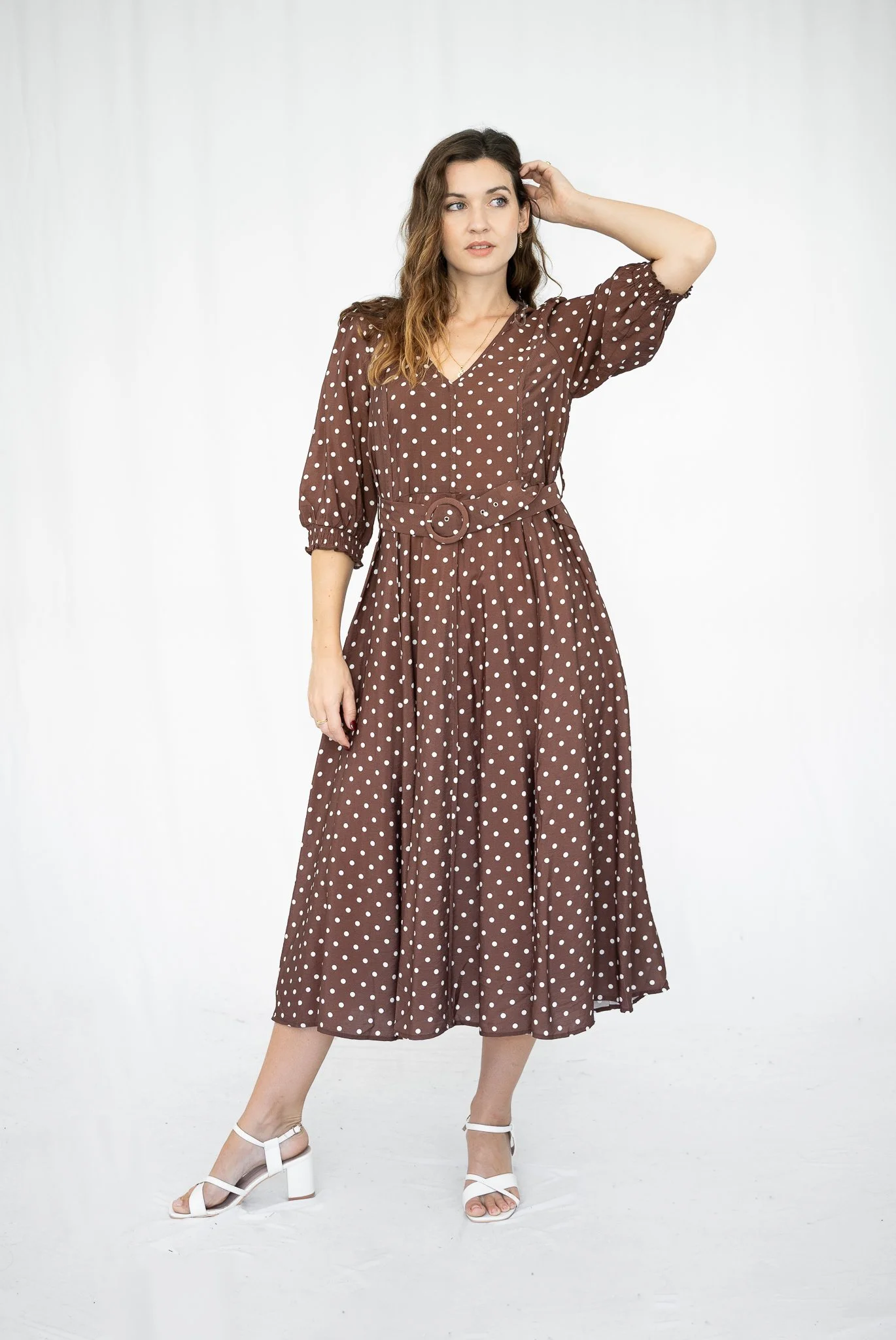 A young woman wearing a brown polka dot dress with puffy sleeves and a belt, white high-heeled sandals, standing against a plain white background, with her right hand touching her hair and her gaze directed to the side.