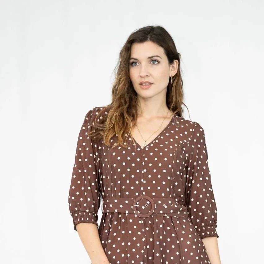 A woman wearing a brown polka dot dress with a belt and ruffled sleeves, standing against a plain white background.