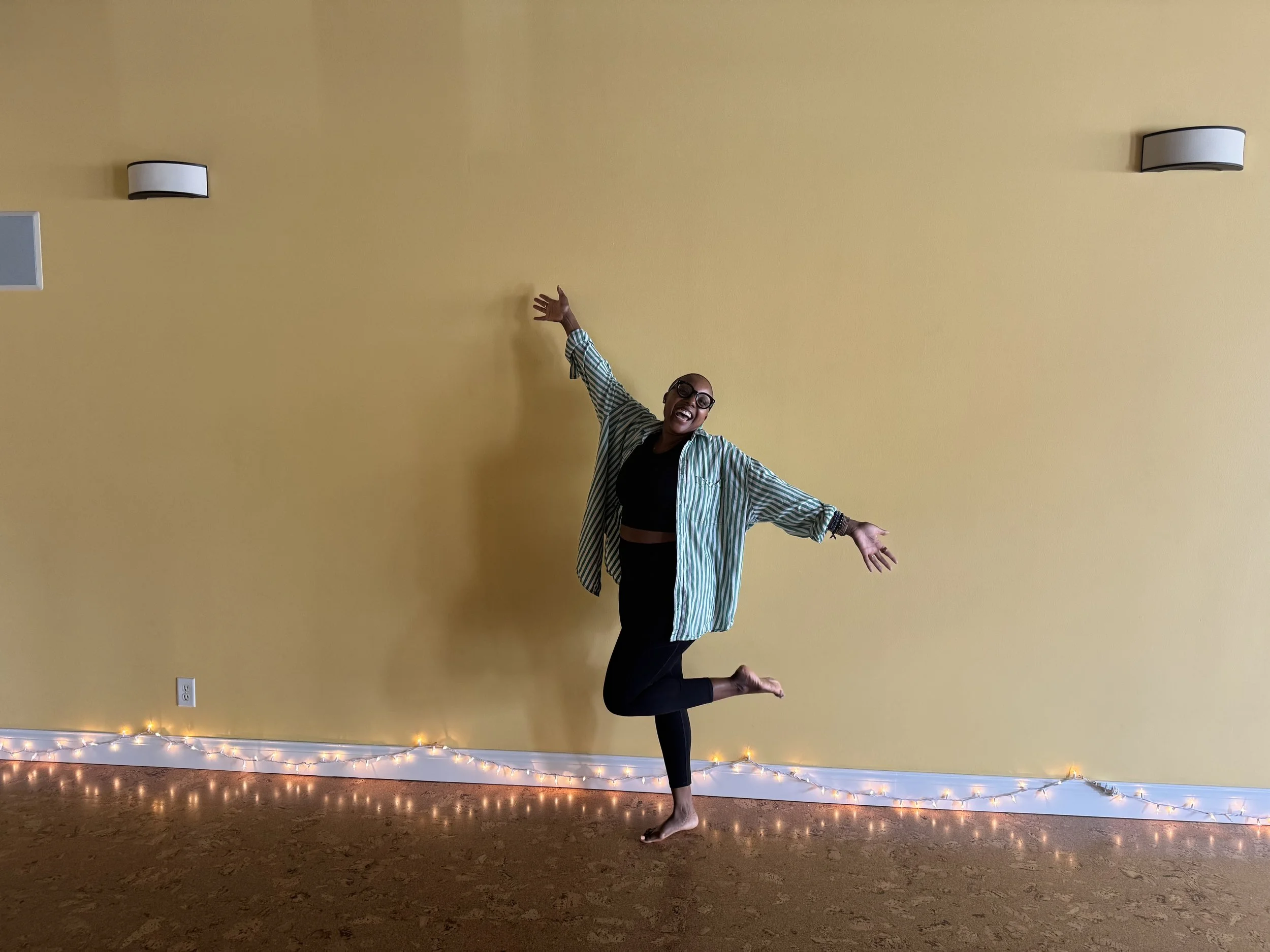 A woman doing a yoga pose on a wooden floor against a yellow wall, with string lights along the baseboard and an electrical outlet. She wears glasses, a black top, black leggings, and a striped shirt, and is smiling with her arms raised.