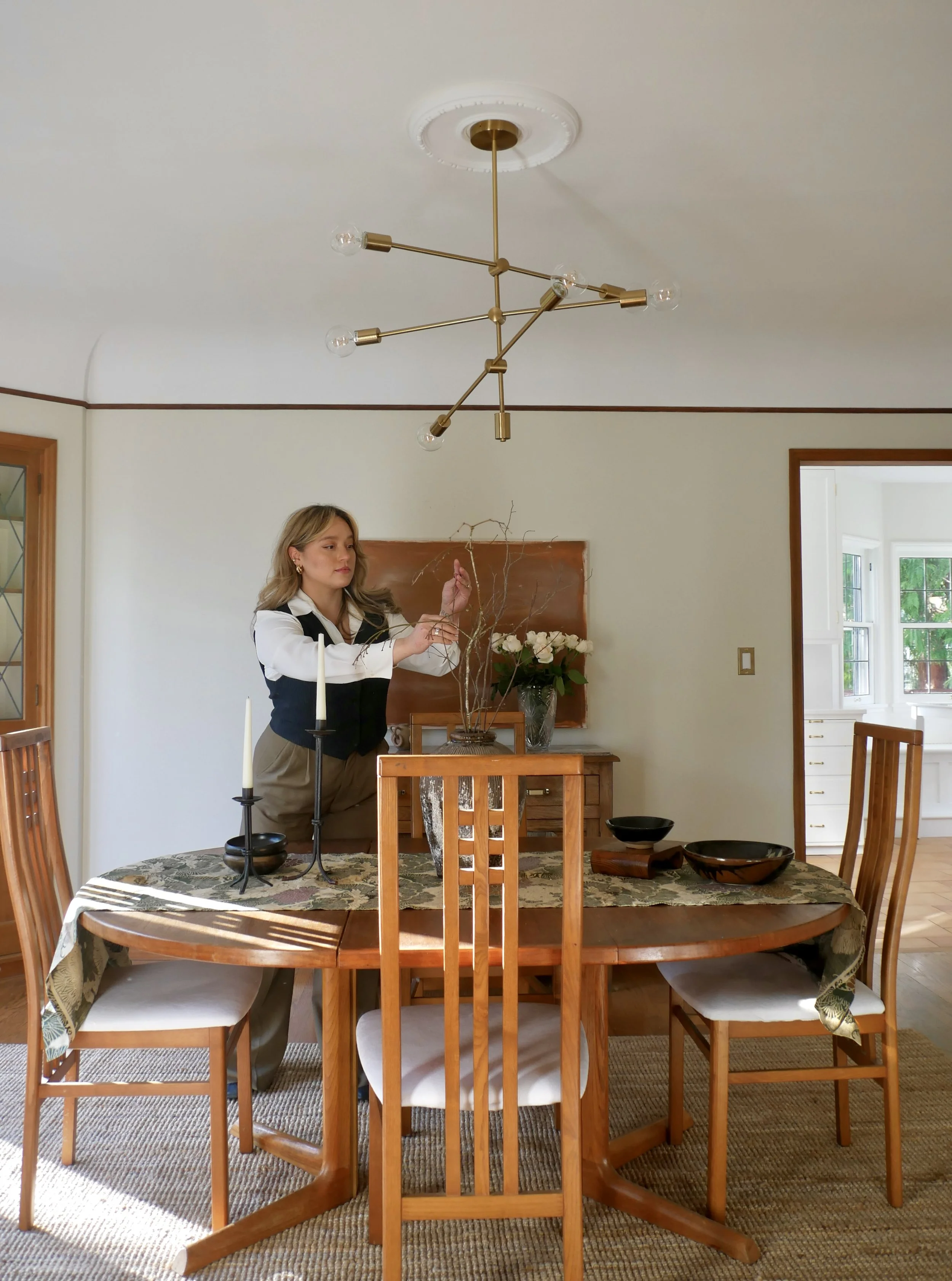 A woman arranging decorative branches on a wooden sideboard in a dining room with a wooden dining table set with black bowls, a candle holder, and a tablecloth. A modern brass chandelier hangs from the ceiling.
