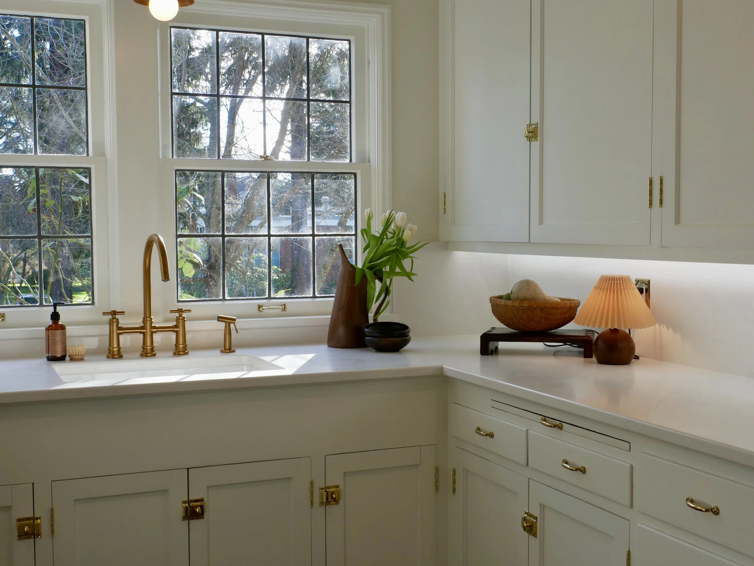 Bright kitchen with white cabinets, gold hardware, and a window view of trees outside. Decor includes a vase of white tulips, a wooden bowl, a small bowl, and an orange table lamp.