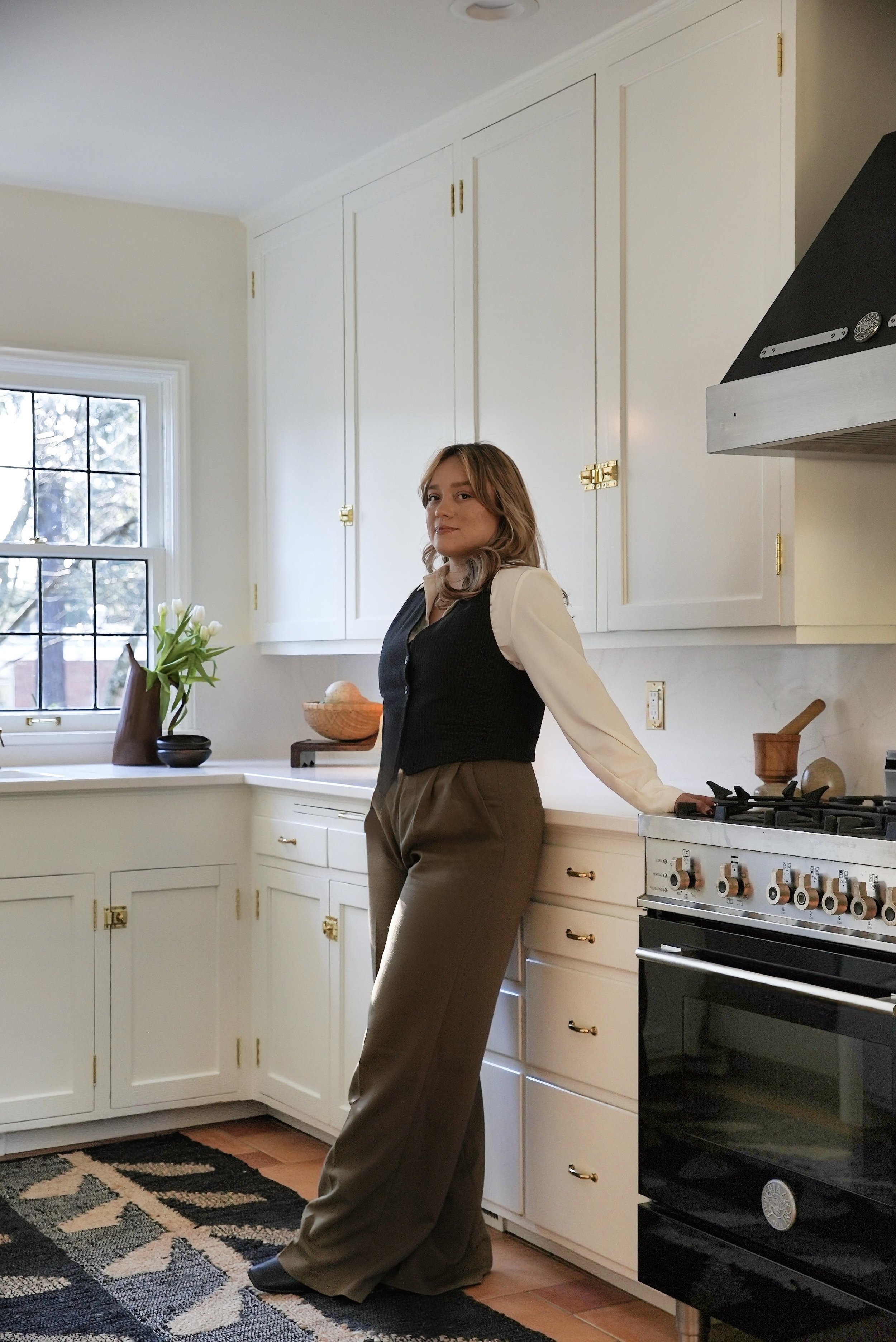A woman standing in a cozy kitchen with white cabinets, a window with black framing, and a black stove, wearing brown pants, a cream blouse, and a black vest.