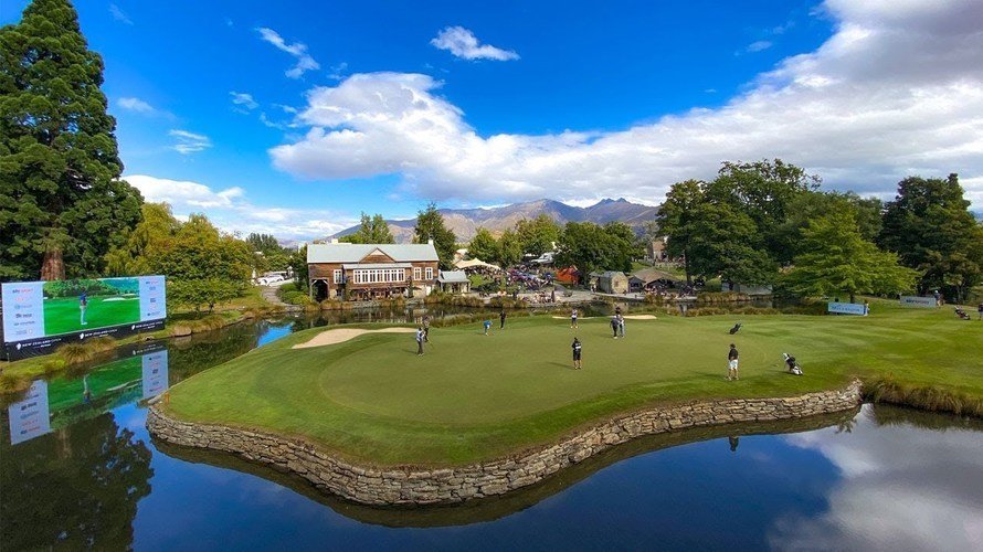 Golf course with players, water hazard, large digital screen, clubhouse, trees, mountains, blue sky with clouds.
