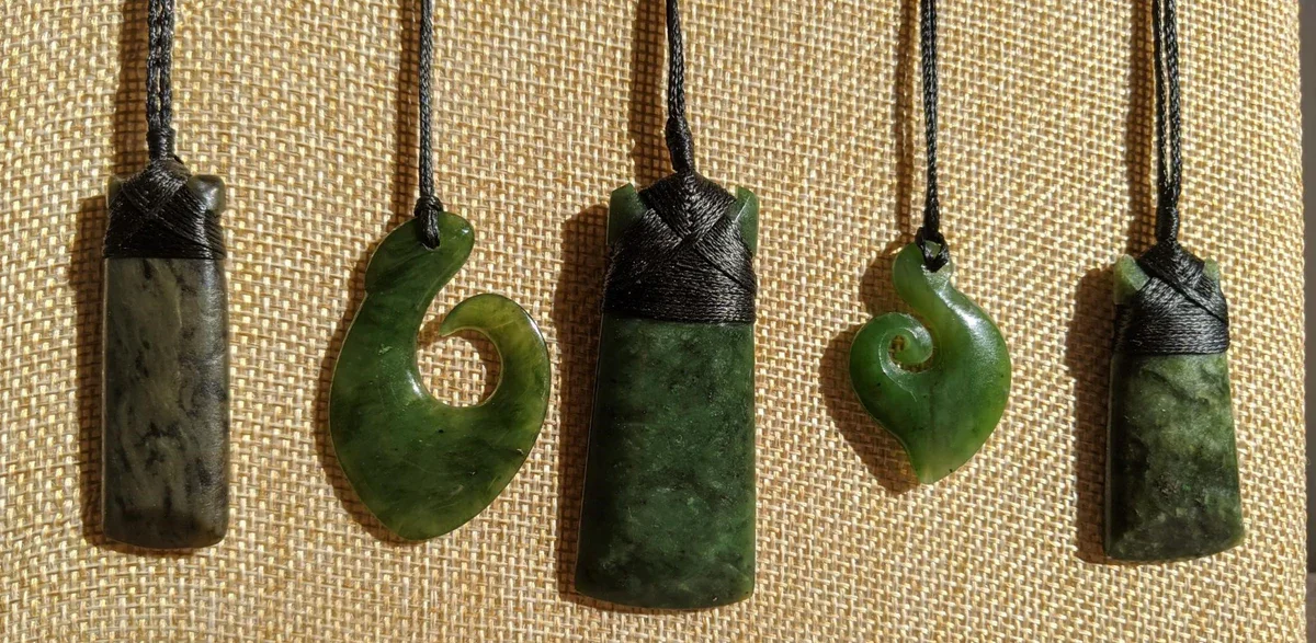 Five green stone pendants on black cords hanging against a beige fabric background.