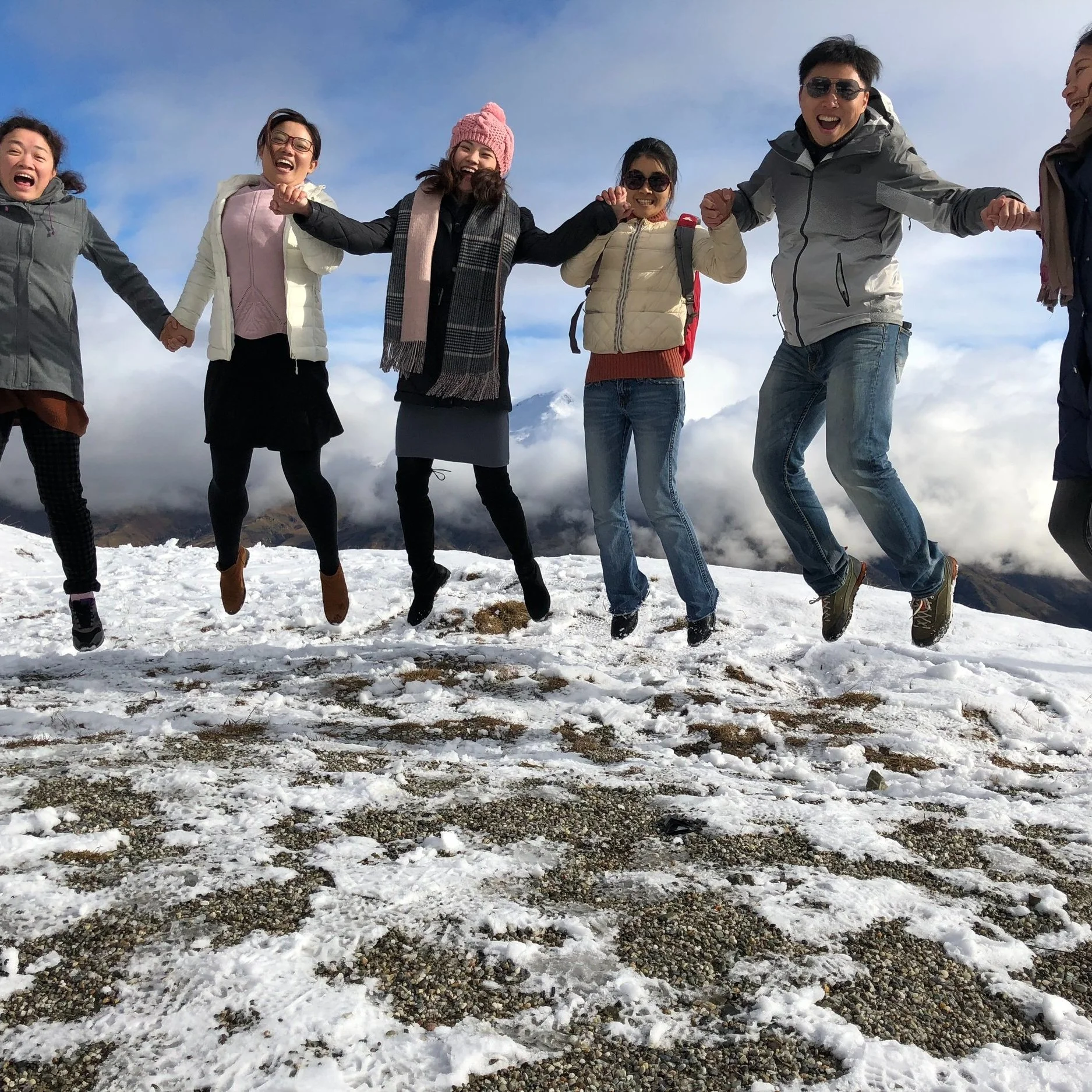 Group of people jumping on snow-covered ground with mountains and clouds in the background, smiling and holding hands.