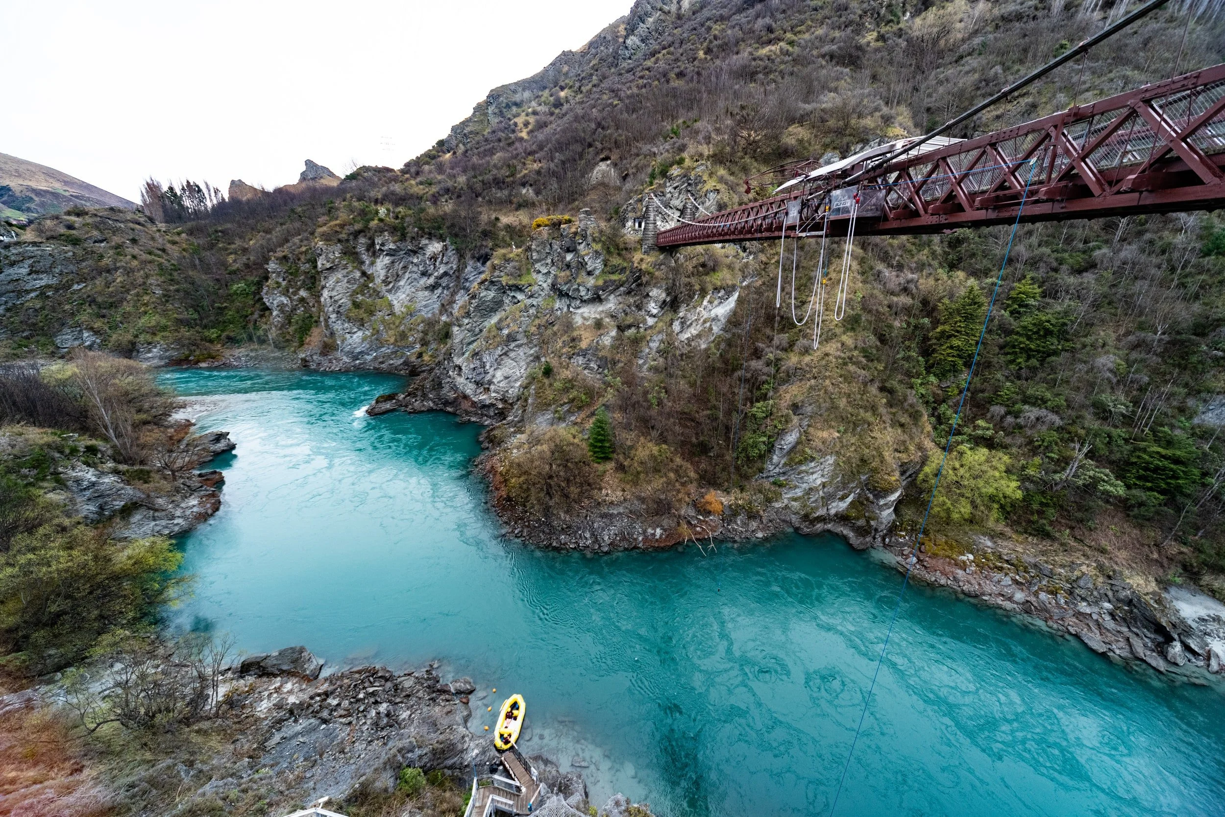 A scenic view of a river with turquoise water flowing through rocky hills, with a narrow red bridge crossing above and a yellow boat docked along the shore.