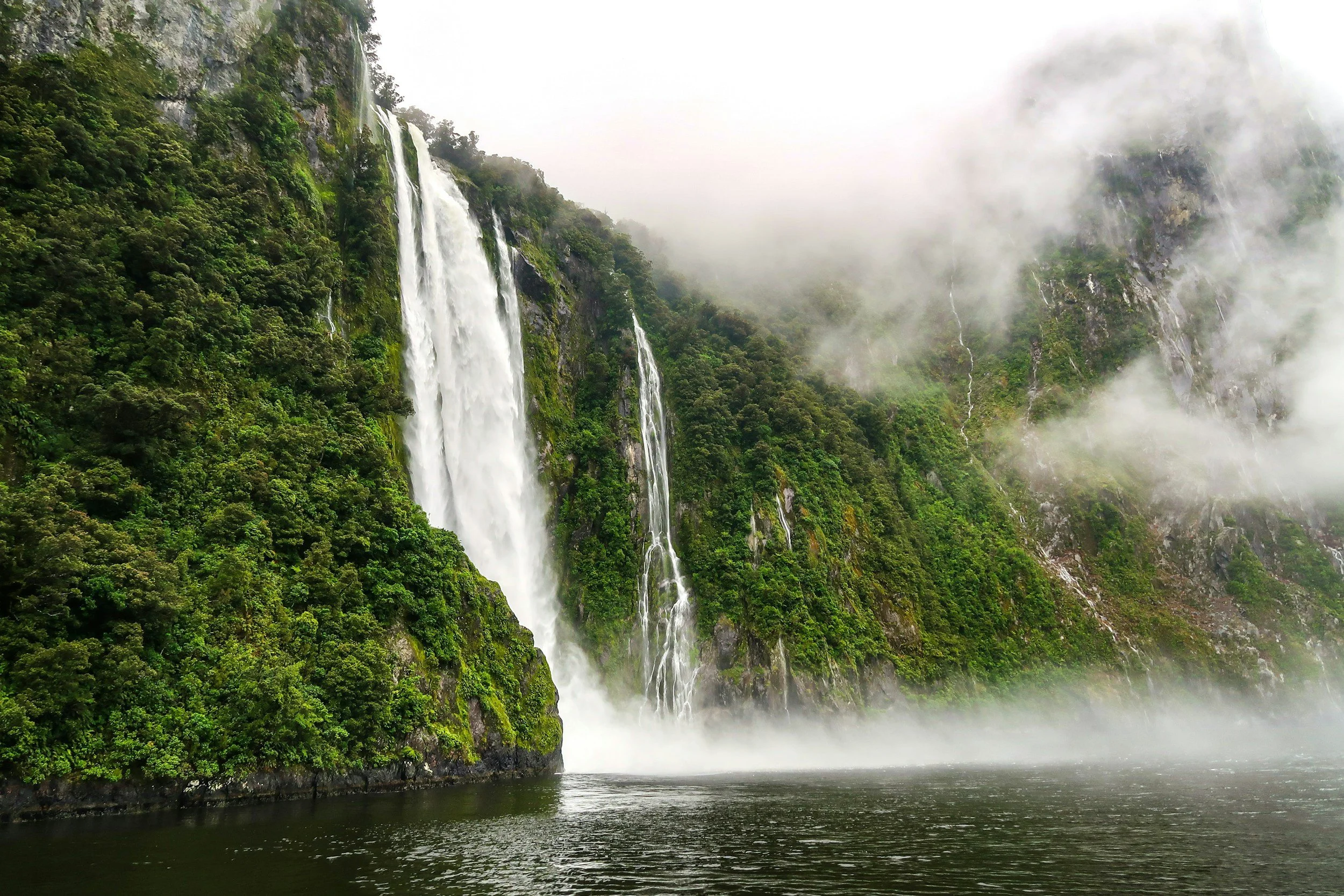 Tall waterfall flowing down lush green mountains with mist and clouds at Milford Sound.
