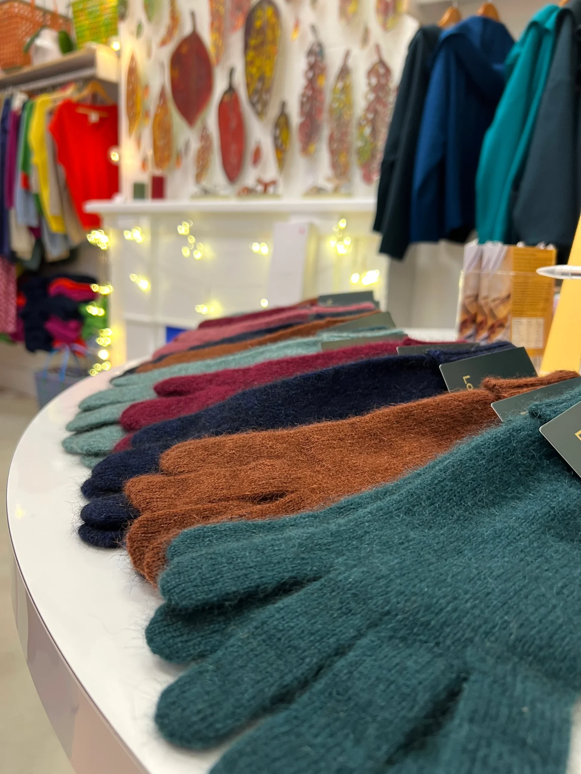 Colorful wool gloves displayed on a table in a store with clothing and artwork in the background.