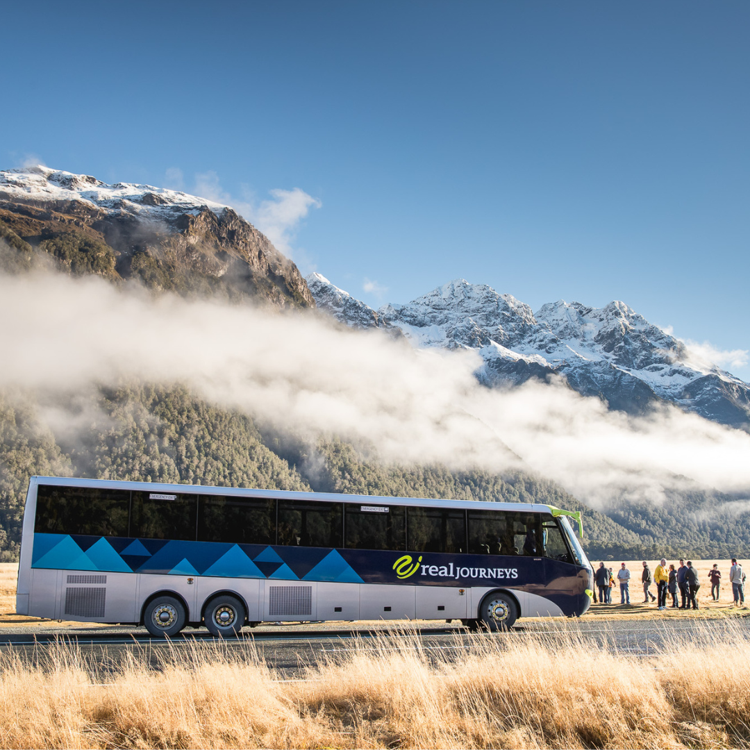 A large tour bus parked on a grassy plain with a mountain range and snow-capped peaks in the background, and a group of people near the bus.