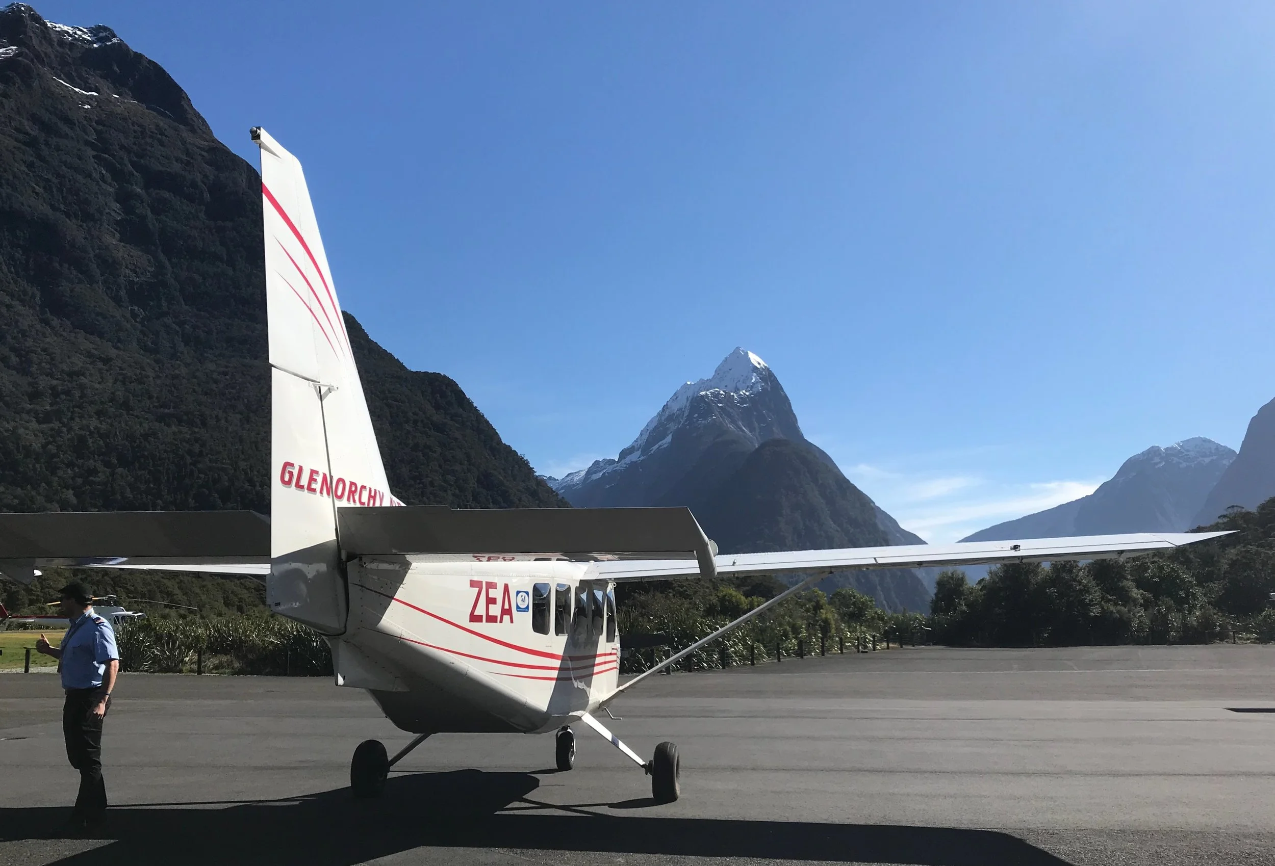 Small white airplane with red markings parked on tarmac near mountainous landscape with snow-capped peaks, a clear blue sky, and trees in the background. A person in a blue shirt and hat is standing near the airplane, looking at their phone.