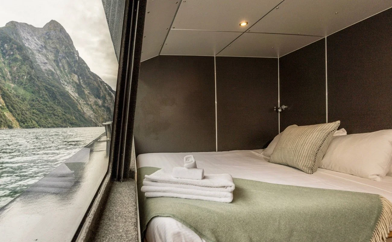 A cozy boat cabin bedroom with a large window showing a waterway and green mountains, neatly made bed with white linens, pillows, a folded green blanket, and rolled towels.