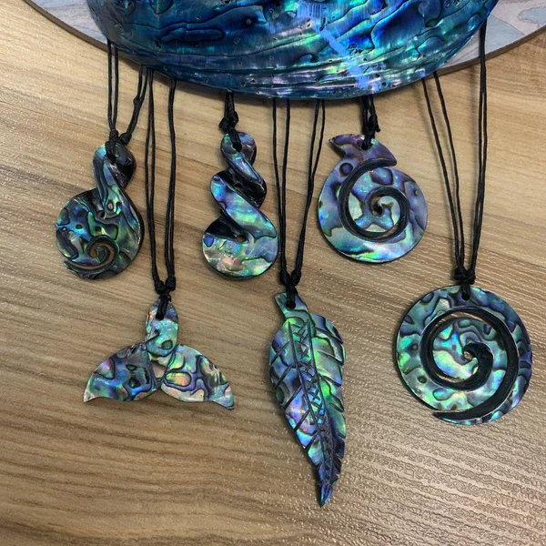 Six shell or abalone jewelry pendants with black cords, including spirals, a whale tail, feather, and leaf shapes, displayed on a wooden surface.