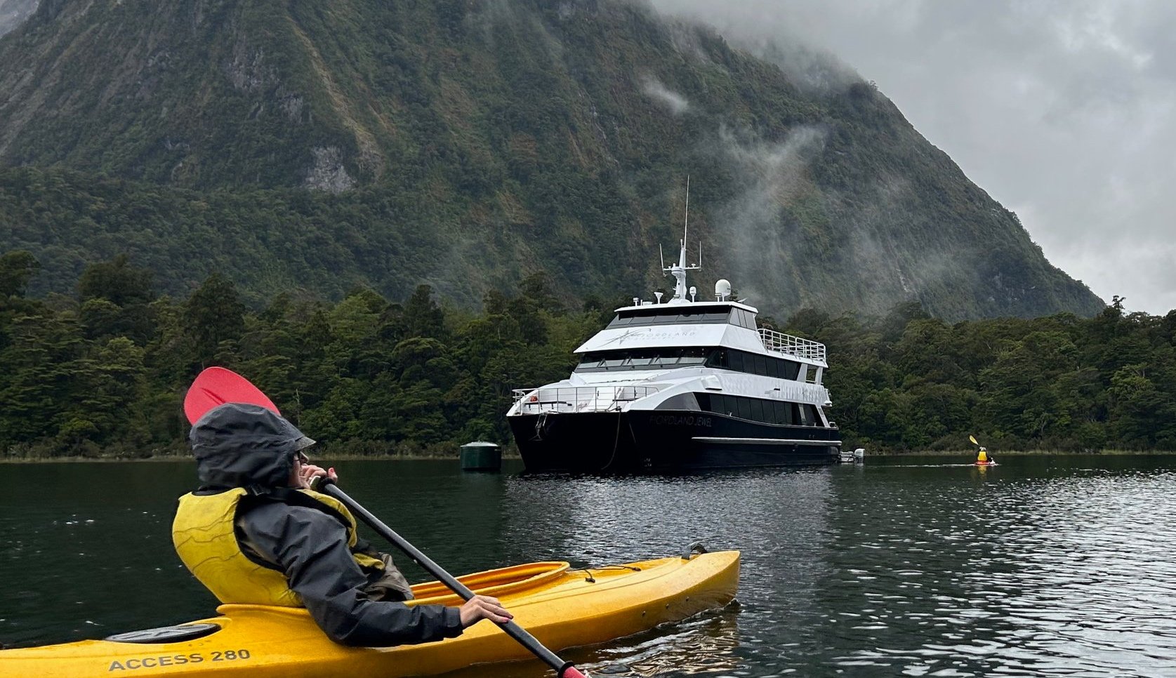 Person in a yellow kayak wearing a rain jacket, paddling near a large white yacht in a body of water surrounded by a green, mountainous landscape with mist and clouds.