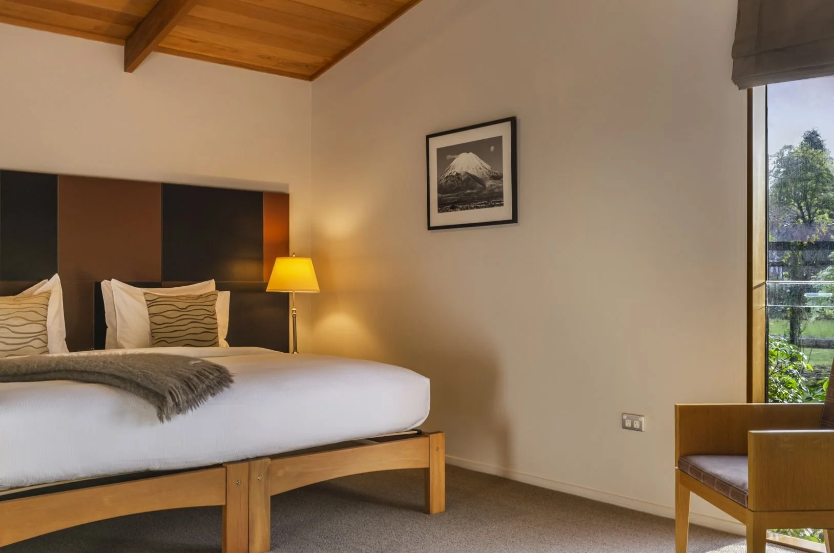 A cozy bedroom with a wooden bed frame, white bedding, and striped pillows, next to a nightstand with a yellow lamp. A framed mountain picture hangs on the wall, and a window shows a view of trees and greenery outside.