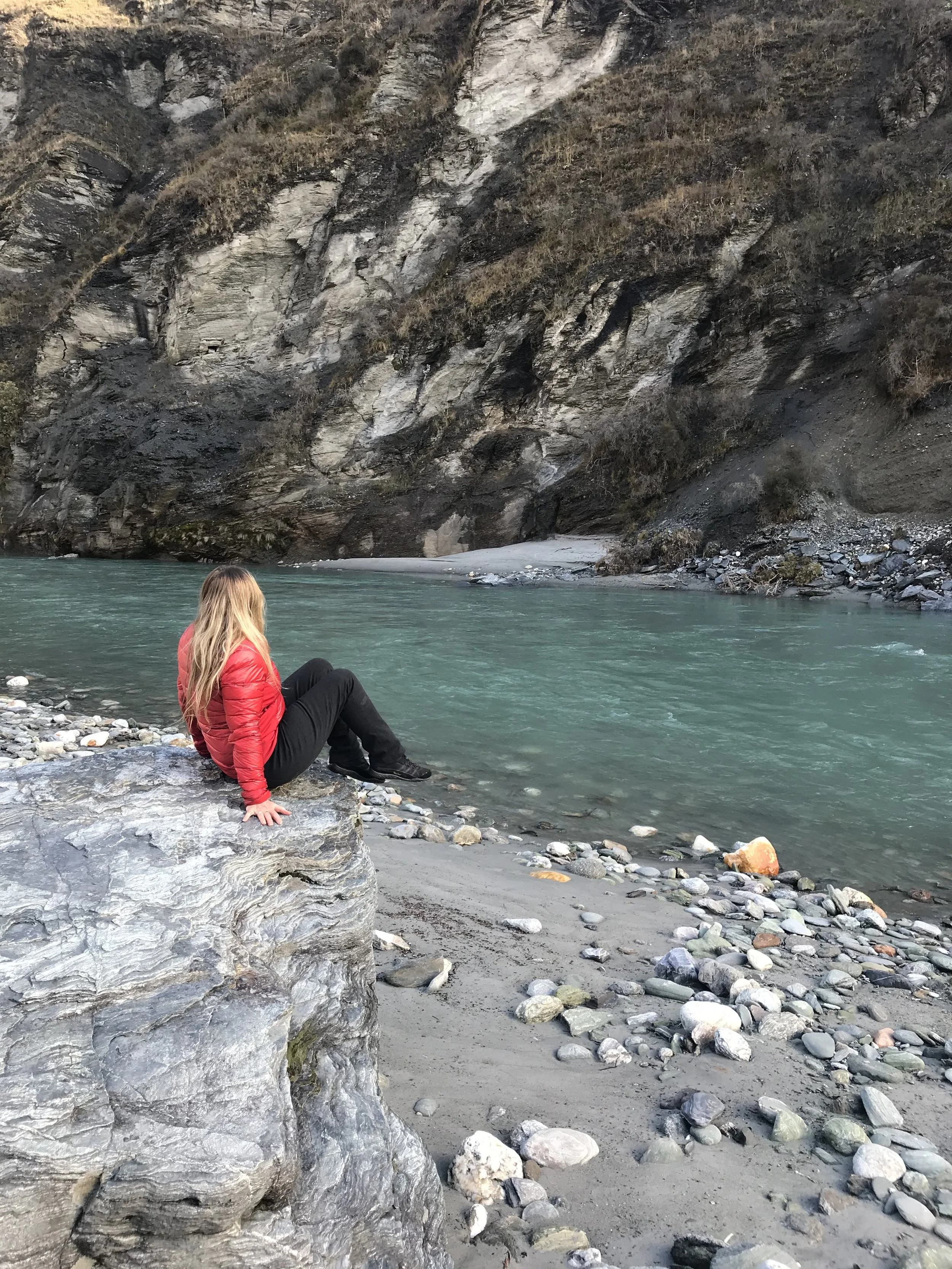A woman with long blonde hair wearing a red jacket and black pants sitting on a large gray rock beside a turquoise river, with steep rocky cliffs in the background.