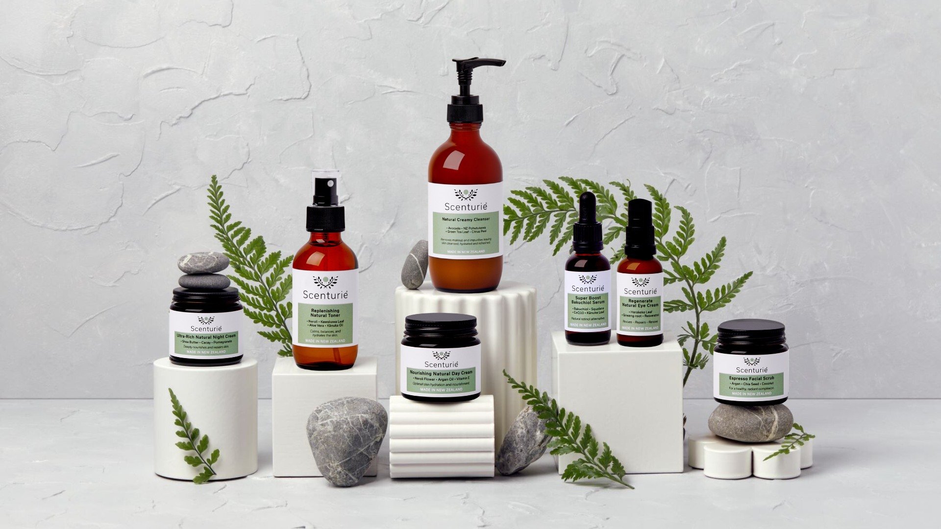 Collection of skincare products from Scenturì, displayed on white pedestals with green fern leaves, gray stones, and a light gray textured background.