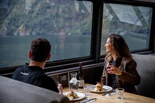A man and a woman dining together on a boat with a scenic water view outside the window, enjoying drinks and food.