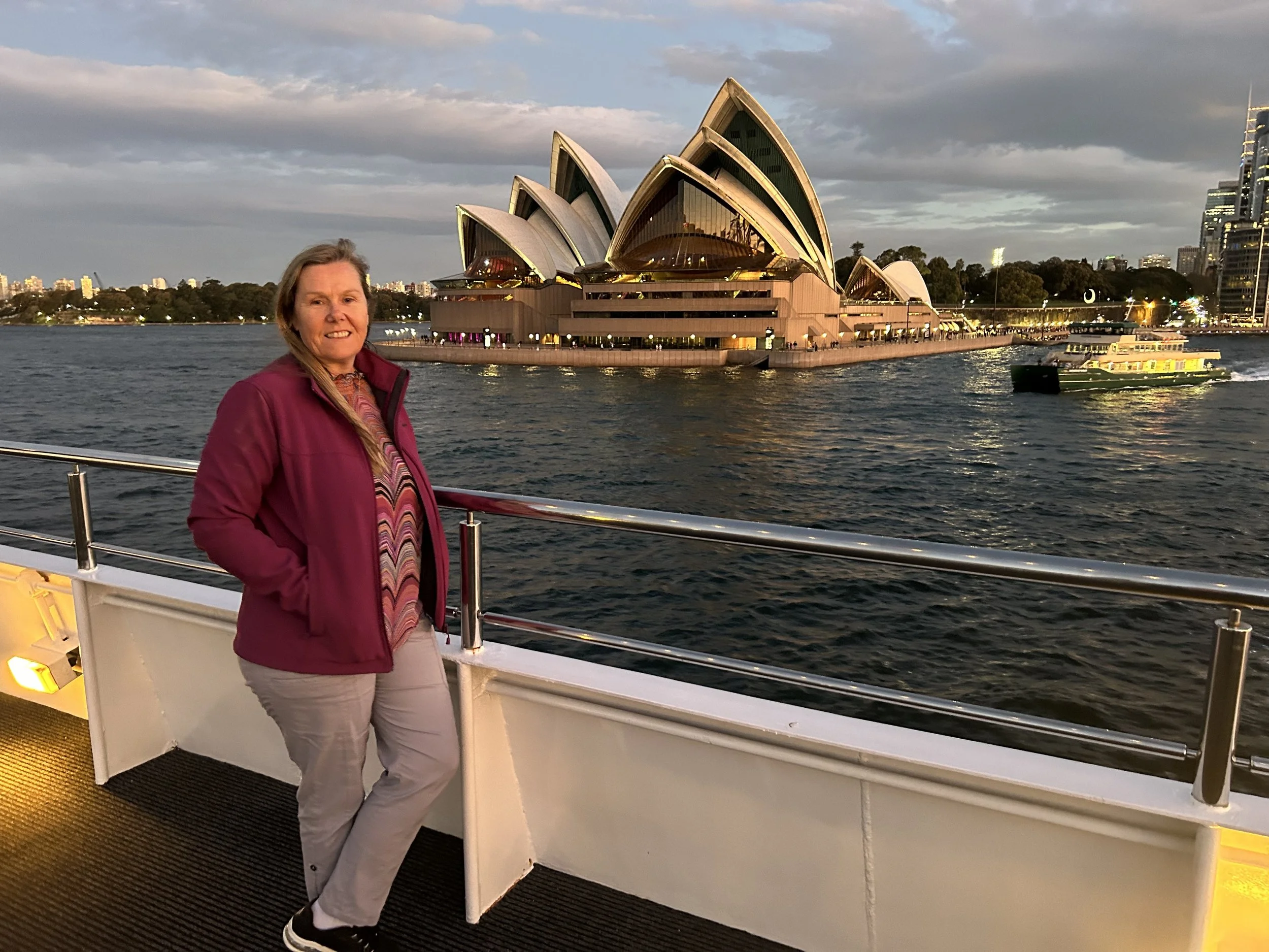 A woman standing on a boat with the Sydney Opera House and city skyline in the background during twilight.
