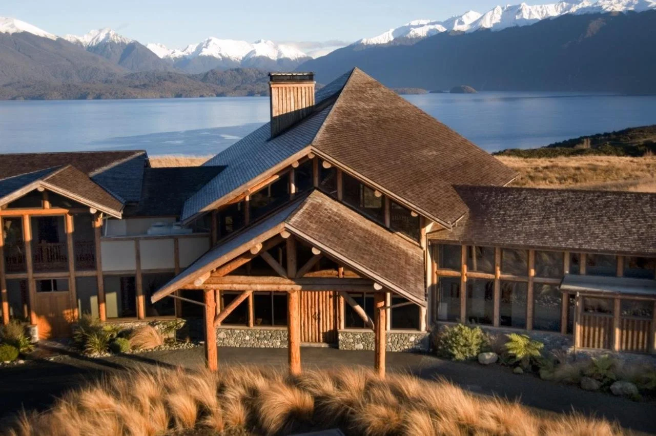 Large wooden lodge with multiple gabled roofs, situated by a lake with snow-capped mountains in the background.
