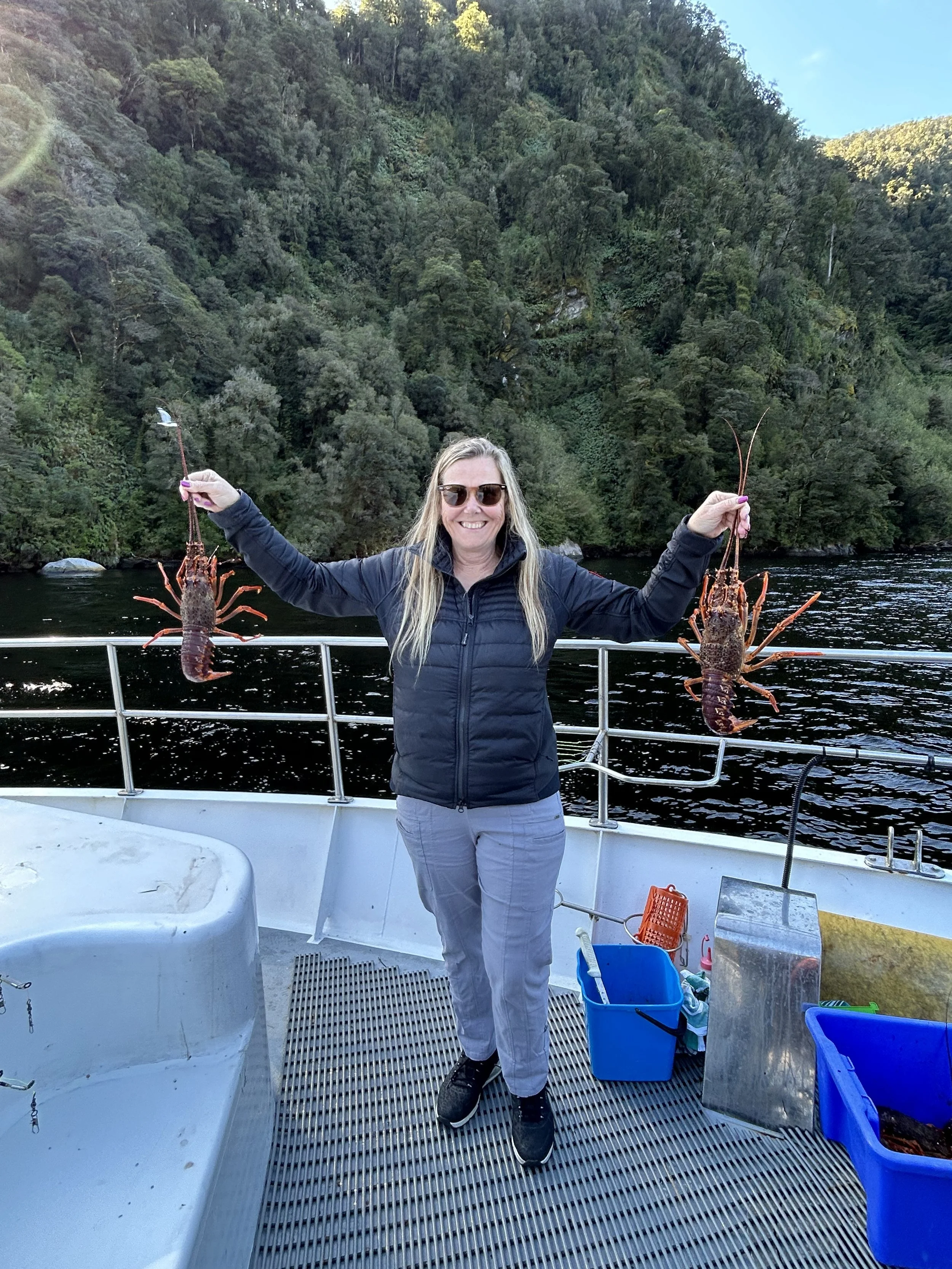 A woman on a boat holding two lobsters, one in each hand, smiling with a backdrop of a forested hillside.