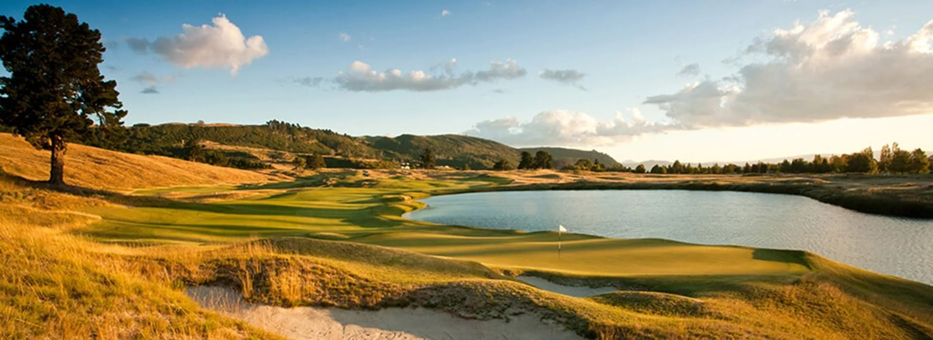 A scenic view of a golf course with a water hazard, green fairways, and sand traps, surrounded by rolling hills under a partly cloudy sky.