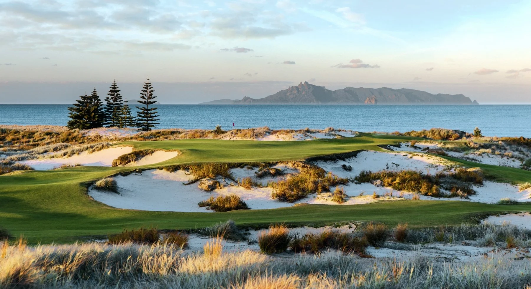Golf course with sand traps and greenery overlooking the ocean, with mountains in the distance and a cloudy sky.