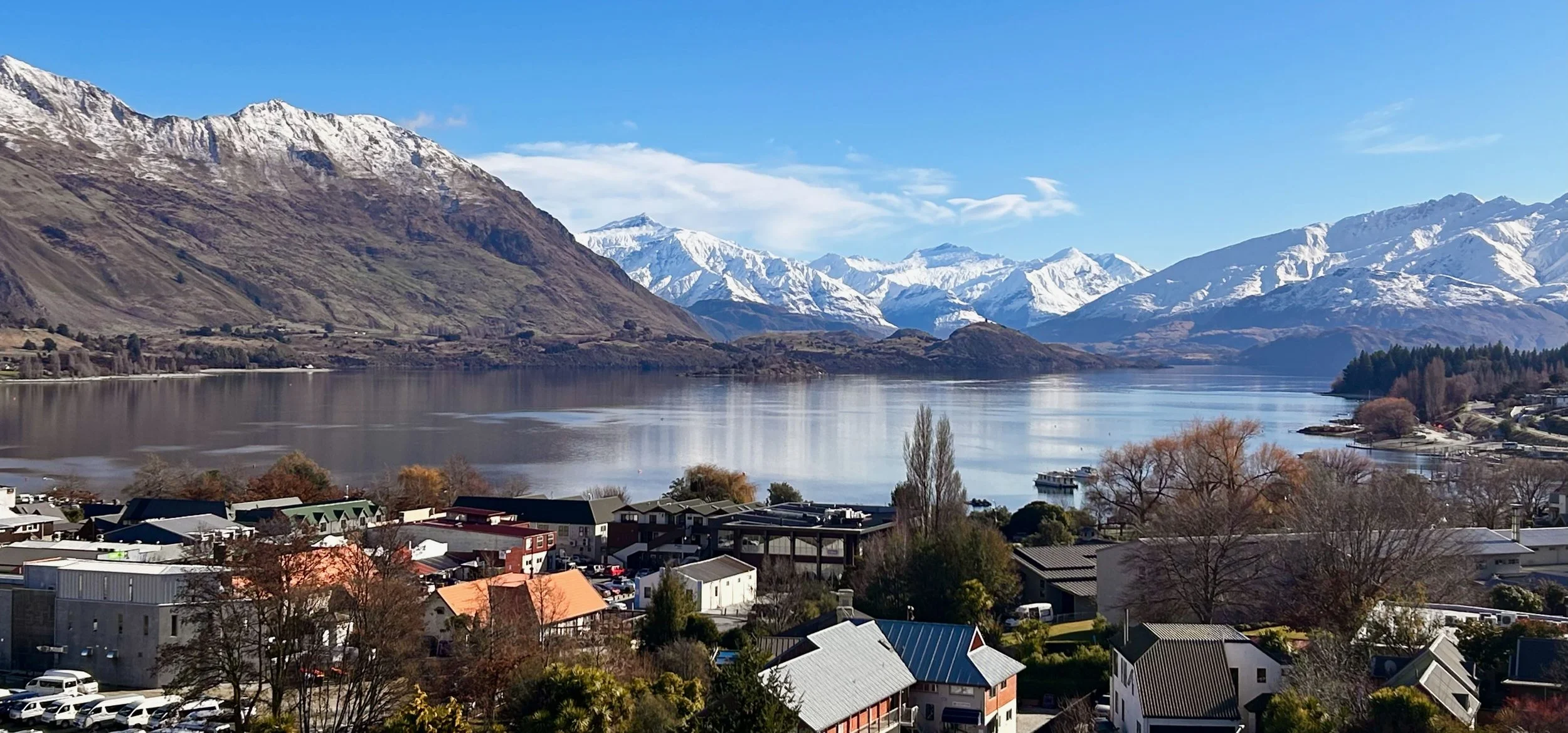 A scenic view of Wānakas lakeside town with snow-capped mountains in the background and a calm lake reflecting the sky.