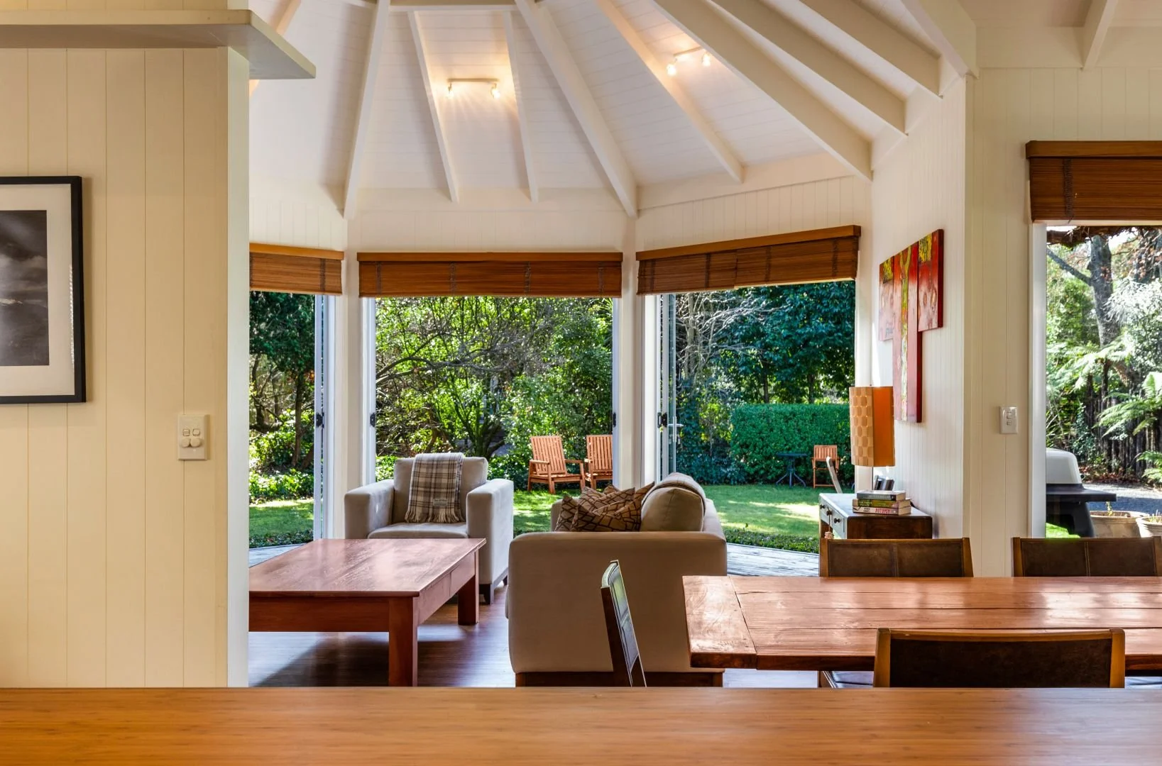 Bright living room with large open windows facing a lush green backyard, with wooden chairs and outdoor scenery.