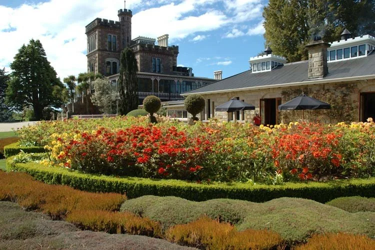 A historic castle-like building with towers and battlements, set behind a landscaped garden with colorful flowers, trimmed hedges, and a house with outdoor umbrellas.