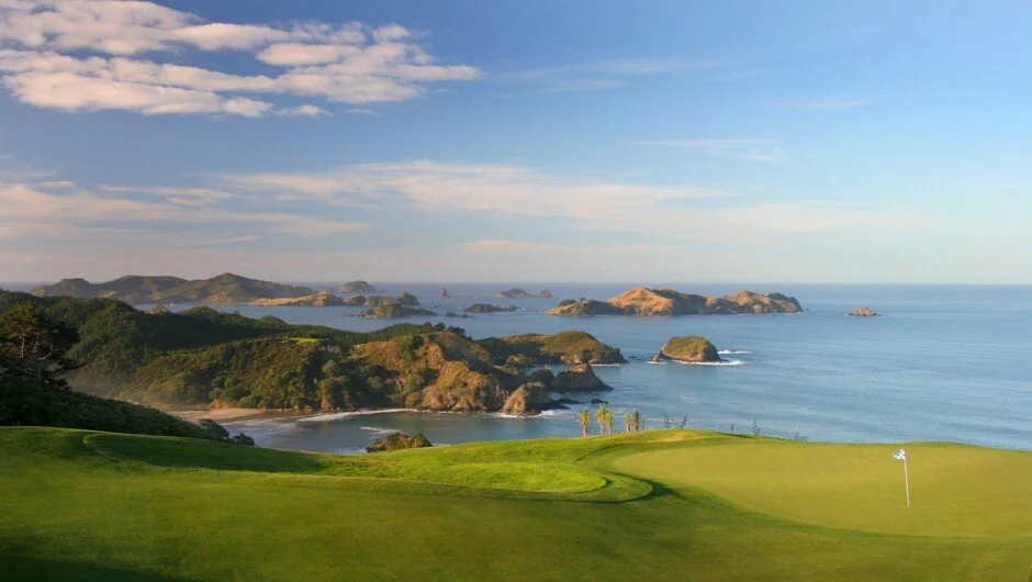 Scenic view of a golf course overlooking a rugged coastline with hills and small islands in the distance under a partly cloudy sky.