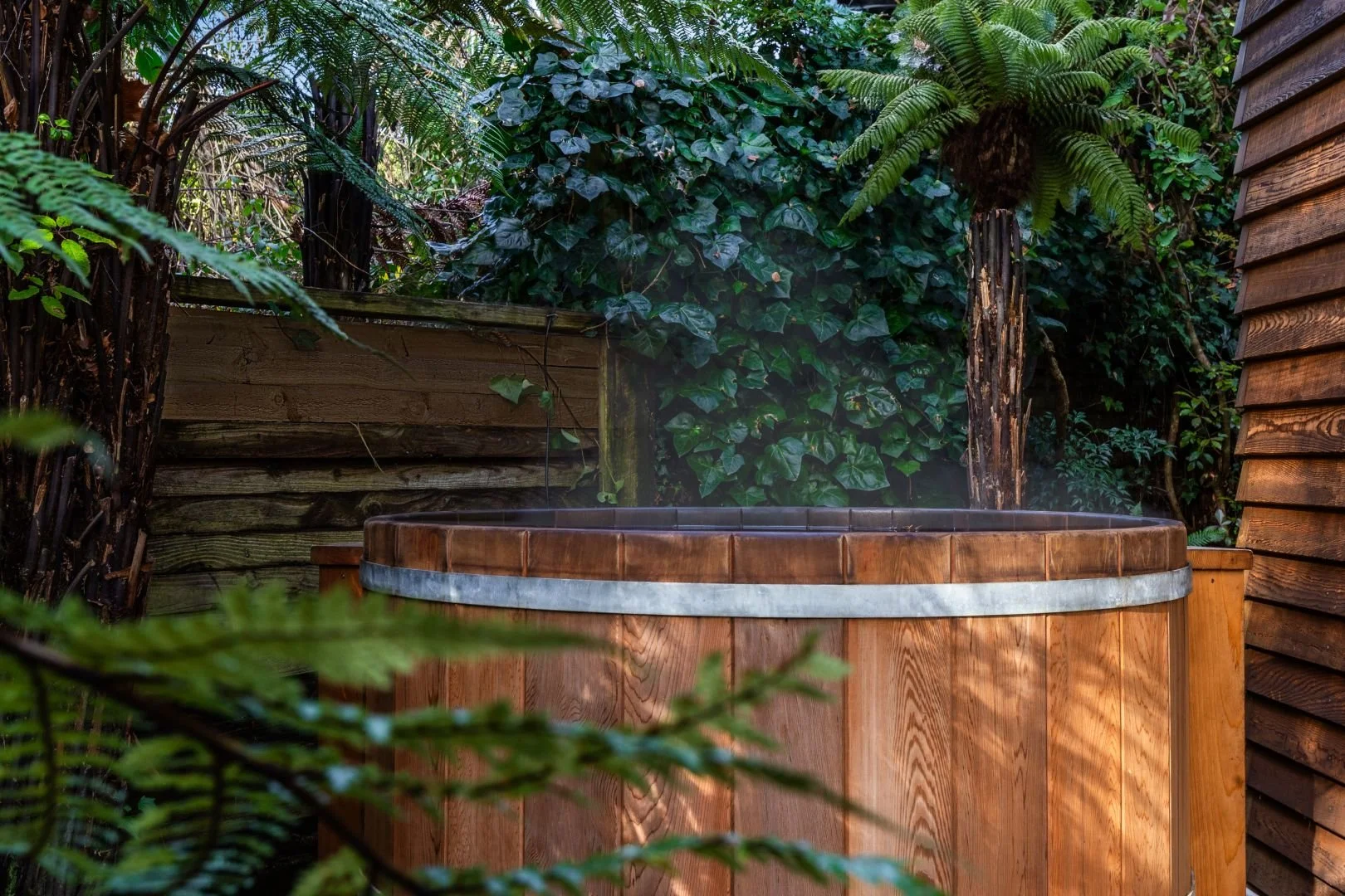 Wooden hot tub on a deck surrounded by lush green plants and foliage.