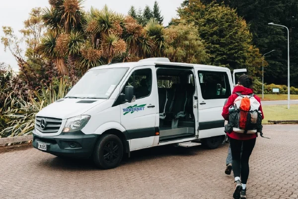 A white shuttle van with an open side door and a person wearing a red jacket and backpack walking towards it, parked on a paved area surrounded by trees and bushes.