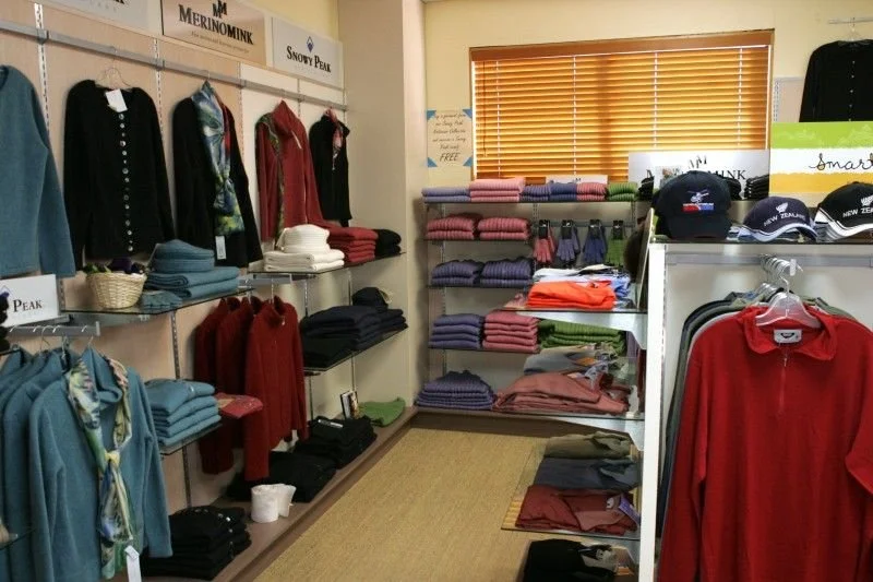 Indoor retail display of clothing including sweaters, hats, and jackets arranged on shelves and racks, with a window with blinds letting in natural light.