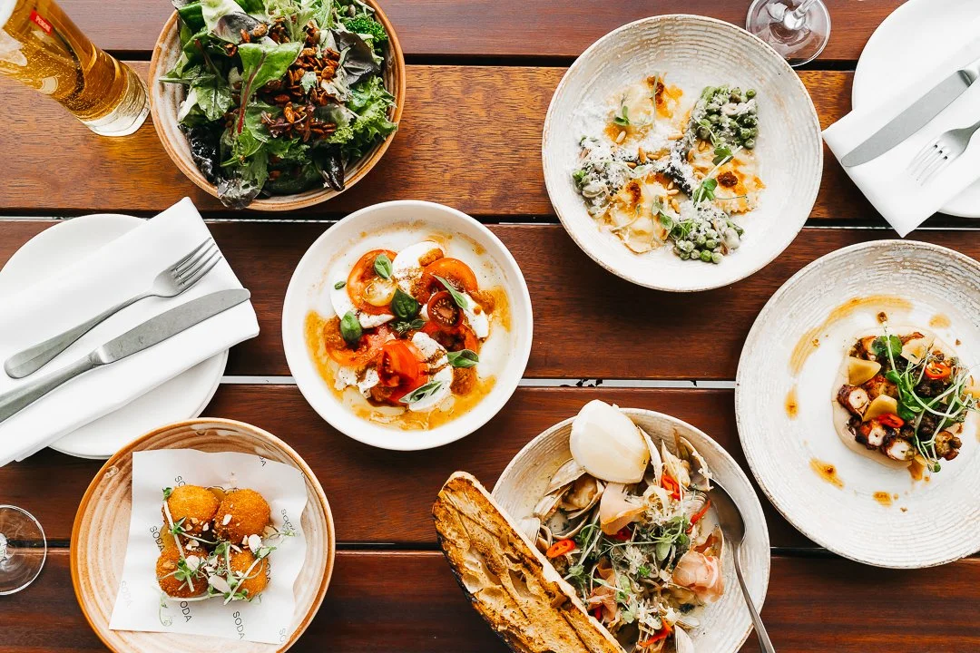 A variety of dishes on a wooden table, including a green salad, a tomato and mozzarella salad, pasta with greens, fried appetizers, seafood, and a bread basket, with utensils and a glass of beer.