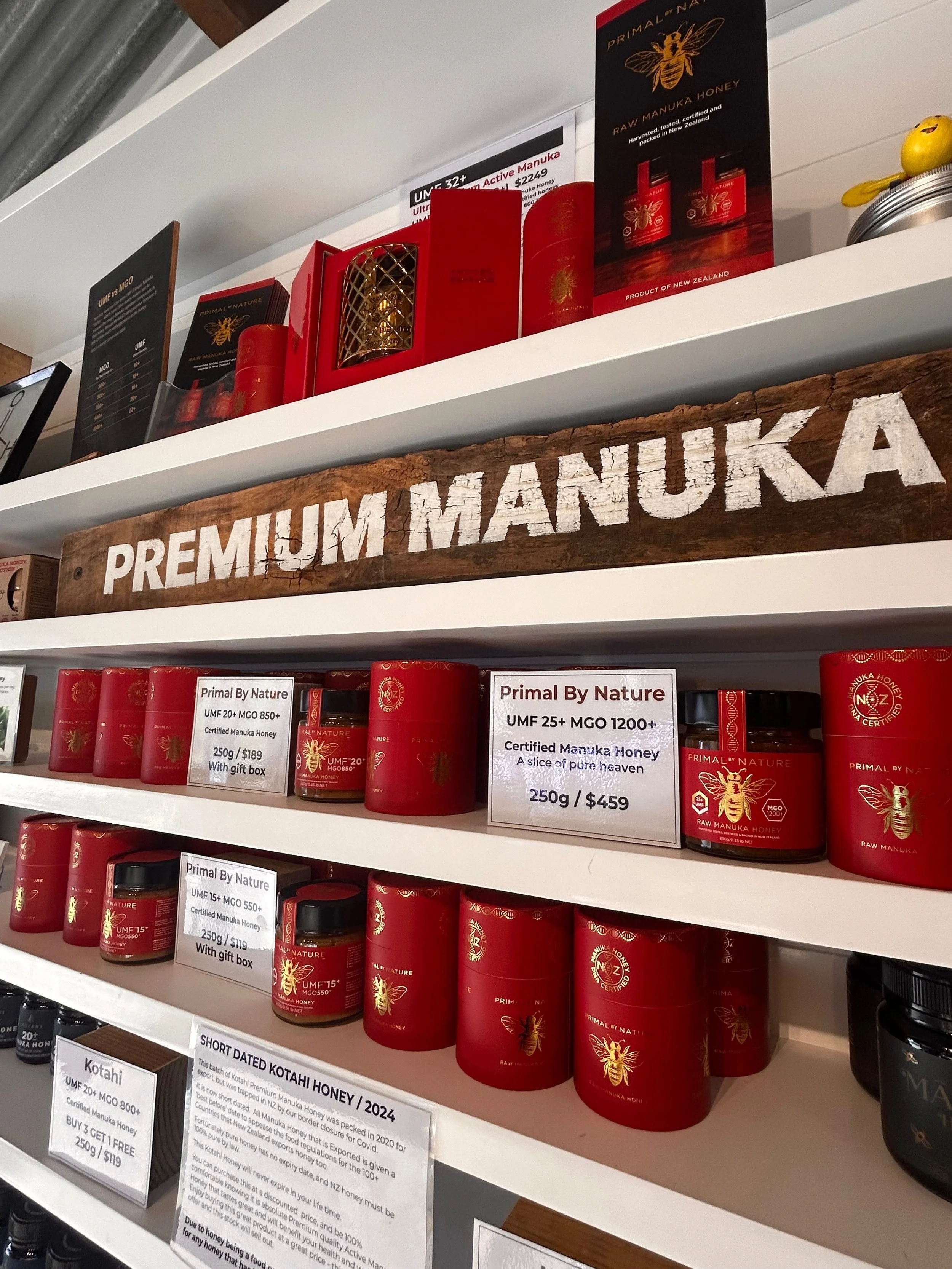 Store shelf displaying various jars and packages of premium Manuka honey in red containers, with signs indicating different grades and prices, and a prominent wooden sign that reads 'PREMIUM MANUKA'.